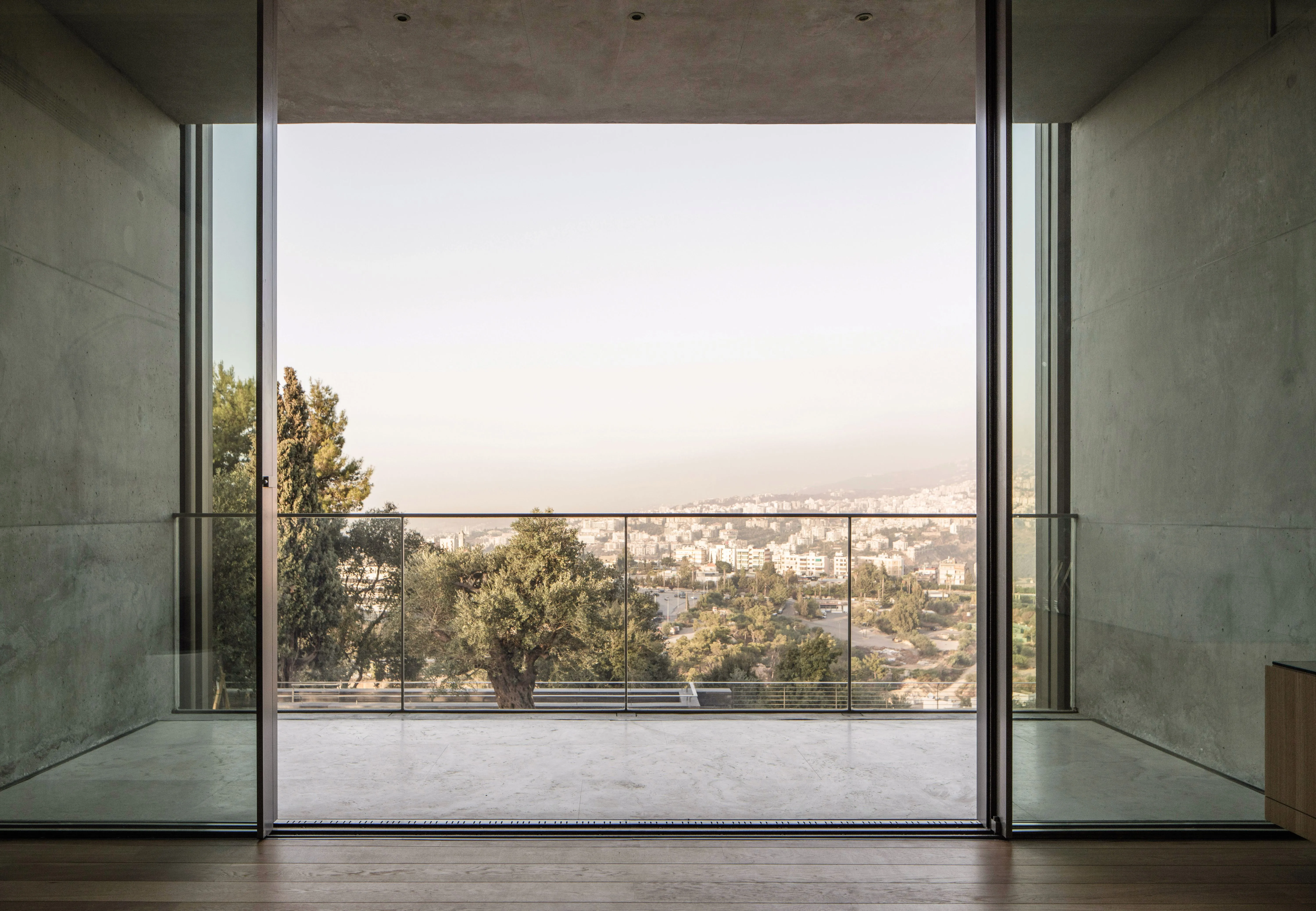 Concrete-framed opening at AY House in Yarze leading to a glass-railed terrace with hillside town views and a mature olive tree below