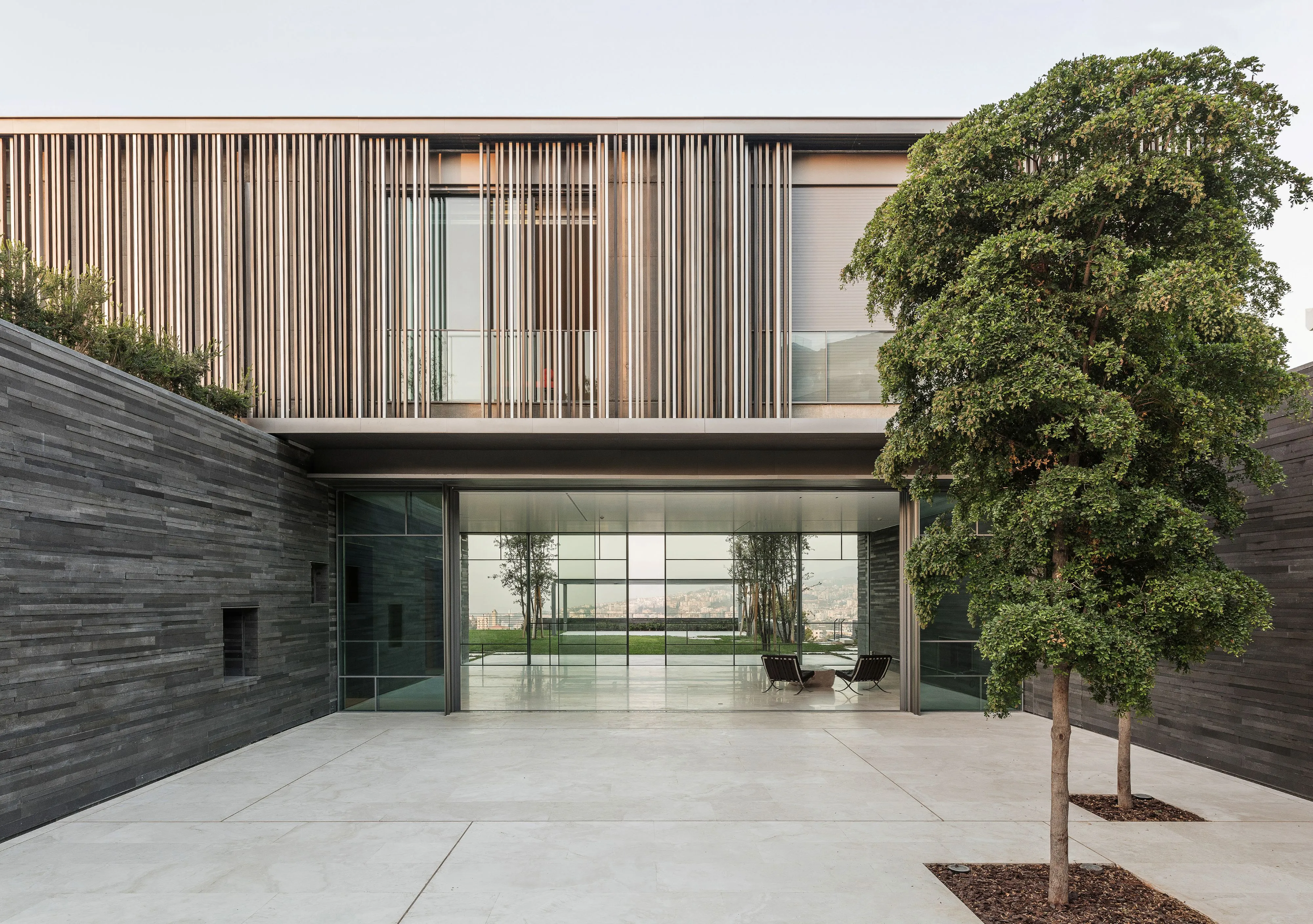 Entrance courtyard of AY House in Yarze, dark basalt walls framing a glazed ground floor that opens to a garden, with timber louvres wrapping the upper storey