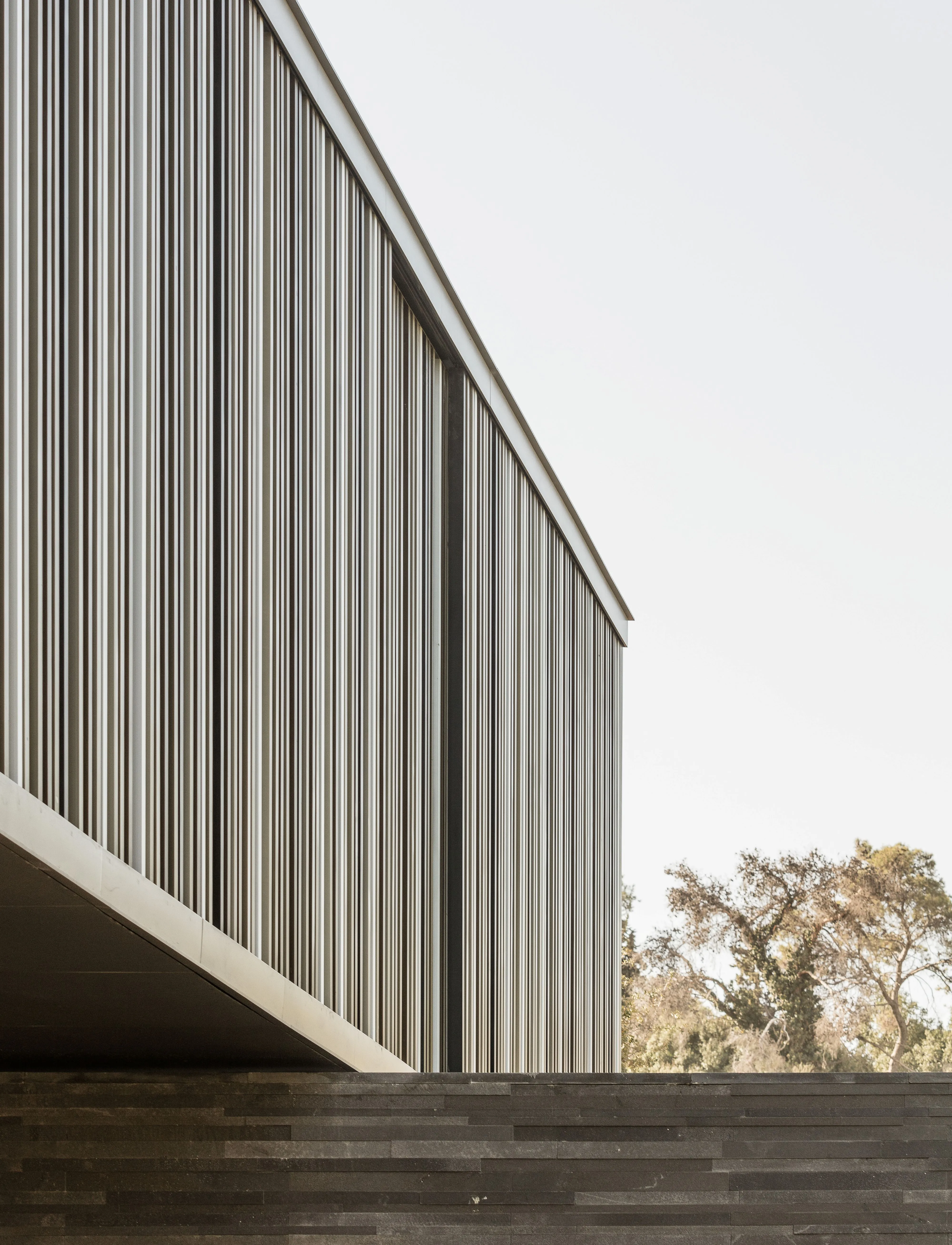 Close-up of AY House in Yarze, vertical louvered cladding on the cantilevered upper level meeting a basalt stone wall, with trees beyond