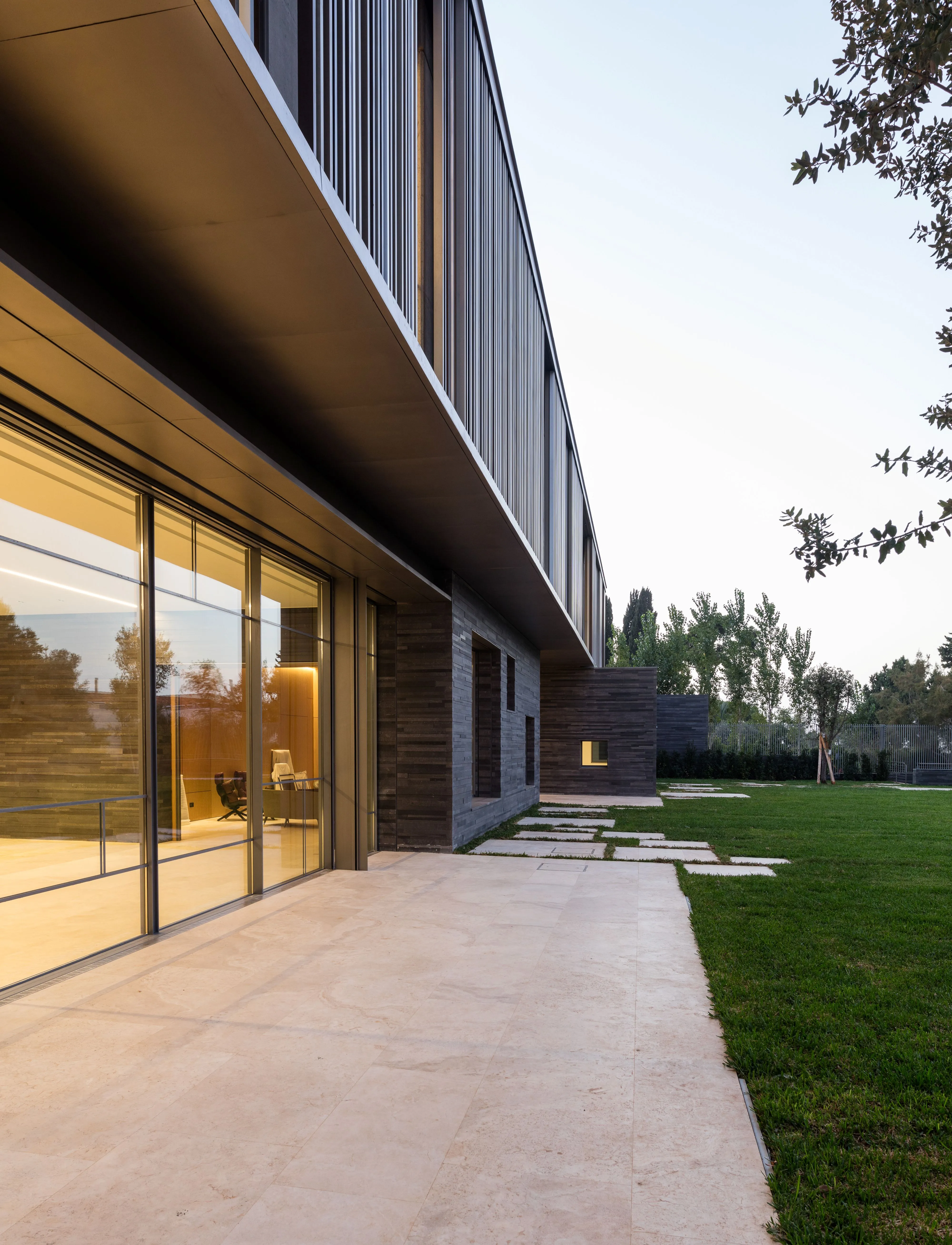 Stone terrace along AY House in Yarze, glazed ground floor glowing warm light beside a lawn, with the louvered upper volume cantilevering above