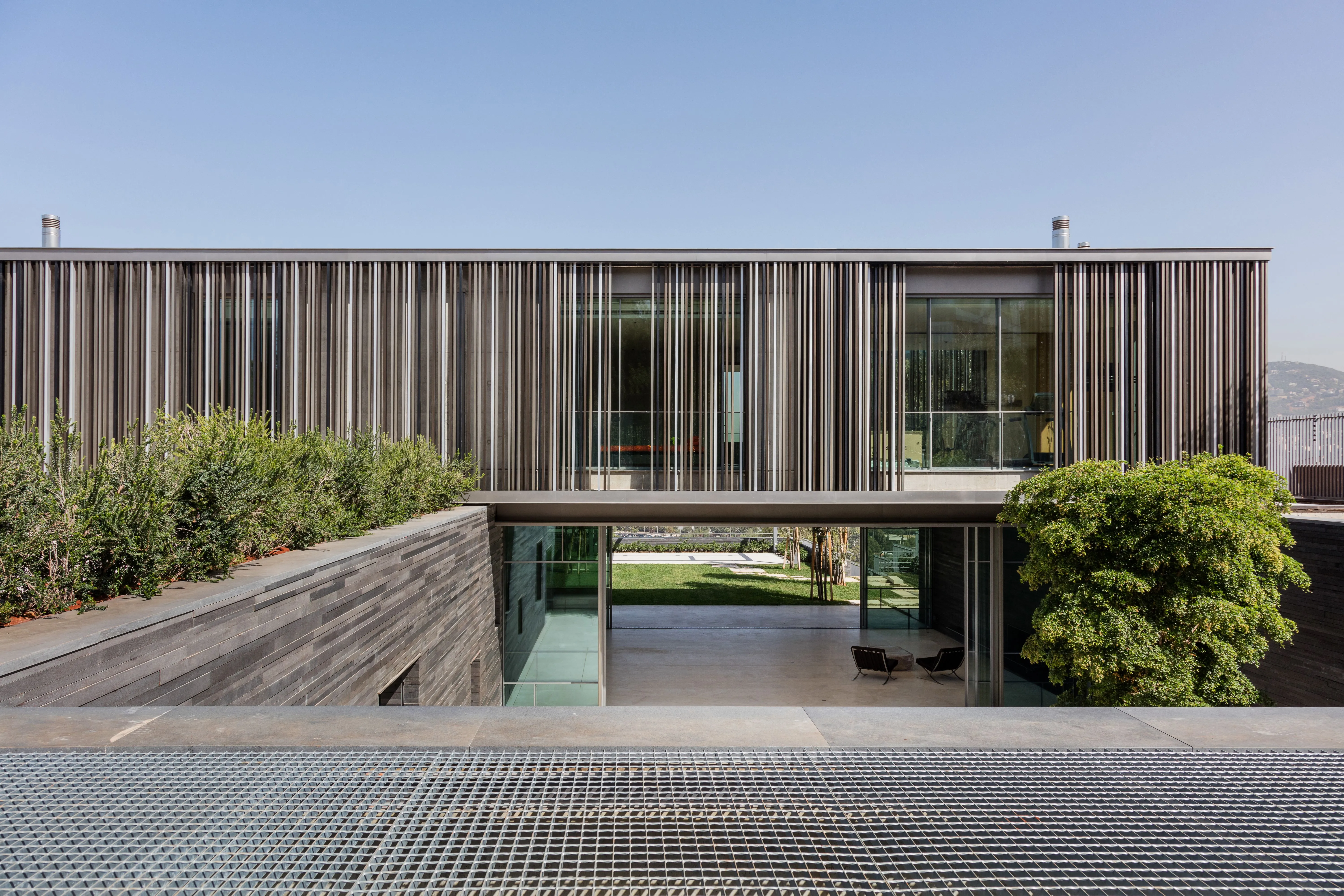 Vertical timber louvers wrap the upper floor of AY House in Yarze, framing a glazed ground level that opens onto a garden and lawn beyond