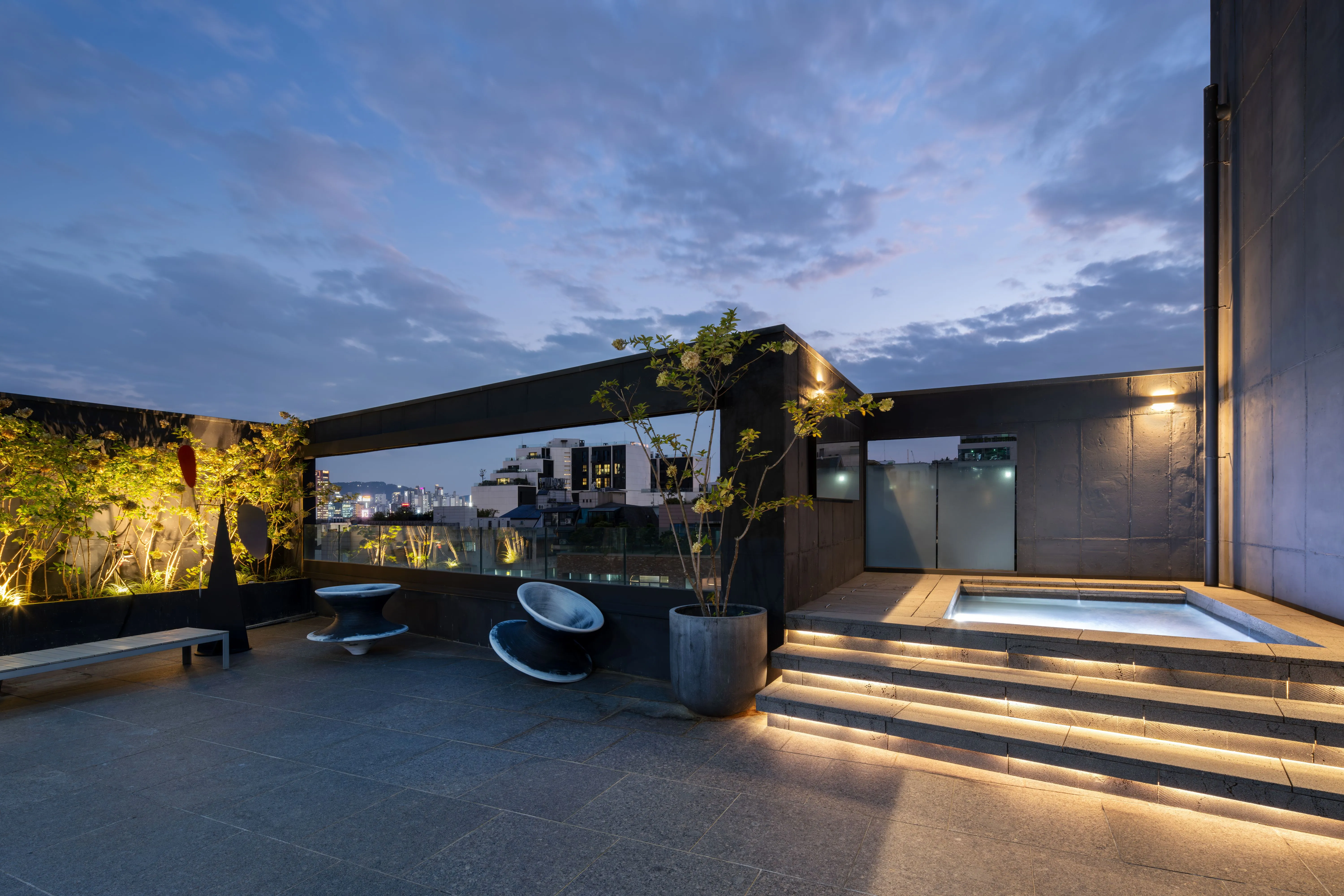 Rooftop terrace of Pyrite in Seoul at dusk, dark concrete walls, illuminated steps leading to a small plunge pool, potted plants and sculptural stone seating