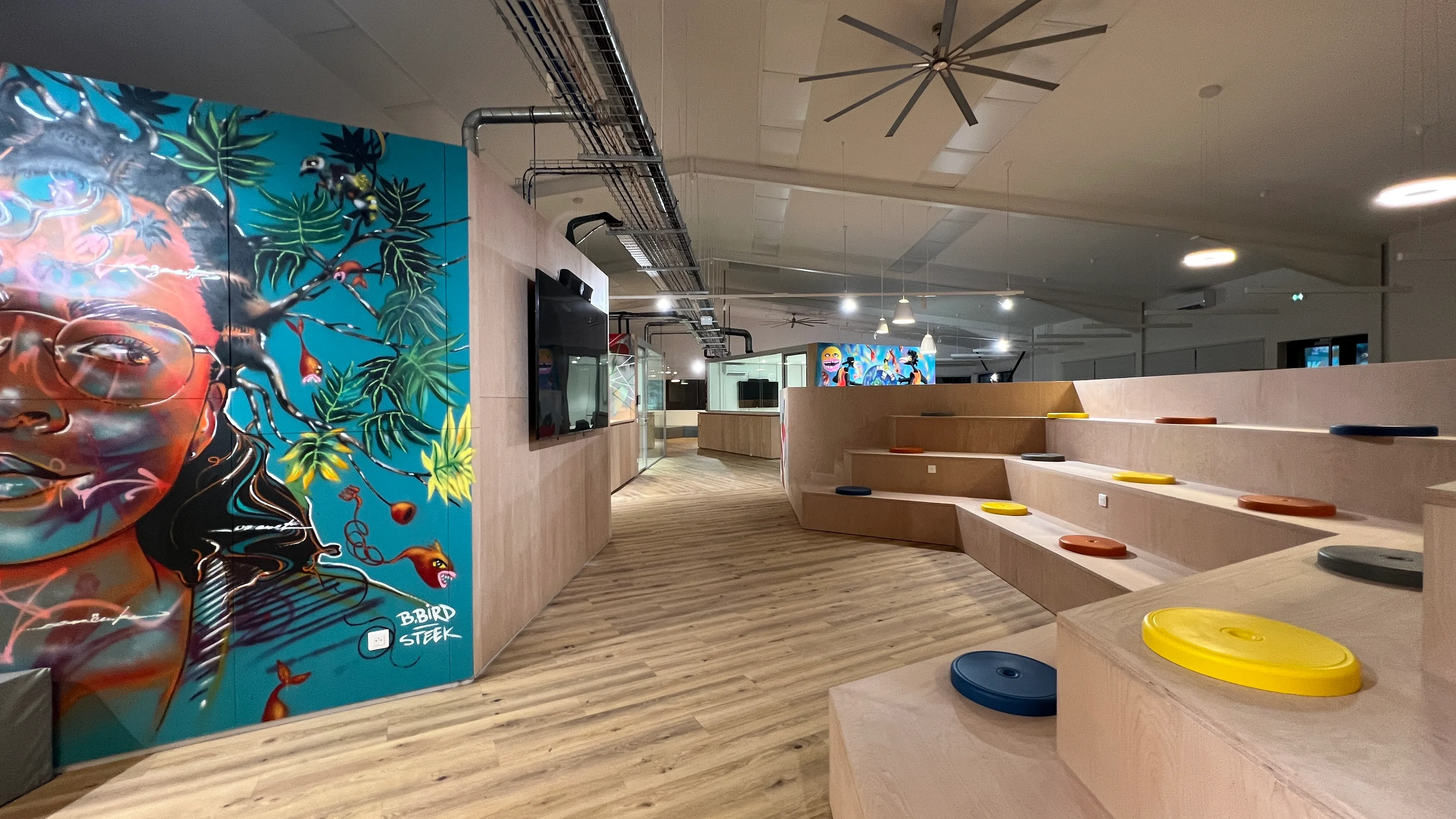 Interior of Zebox Guadeloupe with tiered wooden seating steps, colourful mural of a face and foliage on a blue wall, and exposed ceiling ductwork