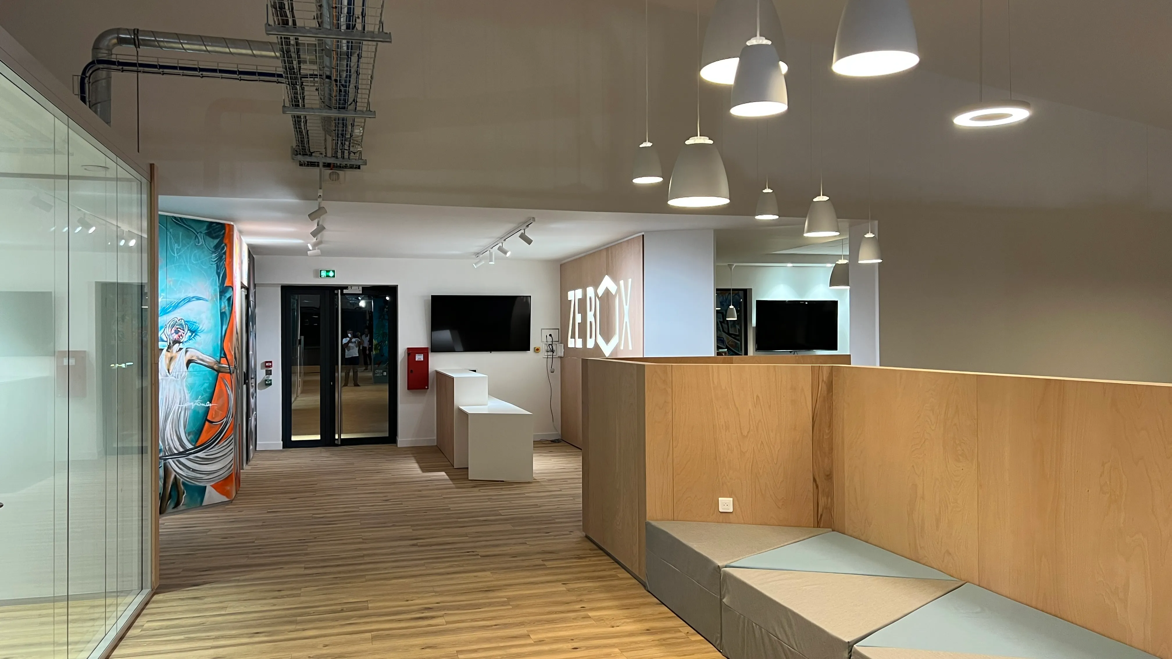 Interior of Zebox Guadeloupe, wooden box-shaped meeting pod with built-in seating beside a wood-floored corridor lit by pendant lamps and a mural wall