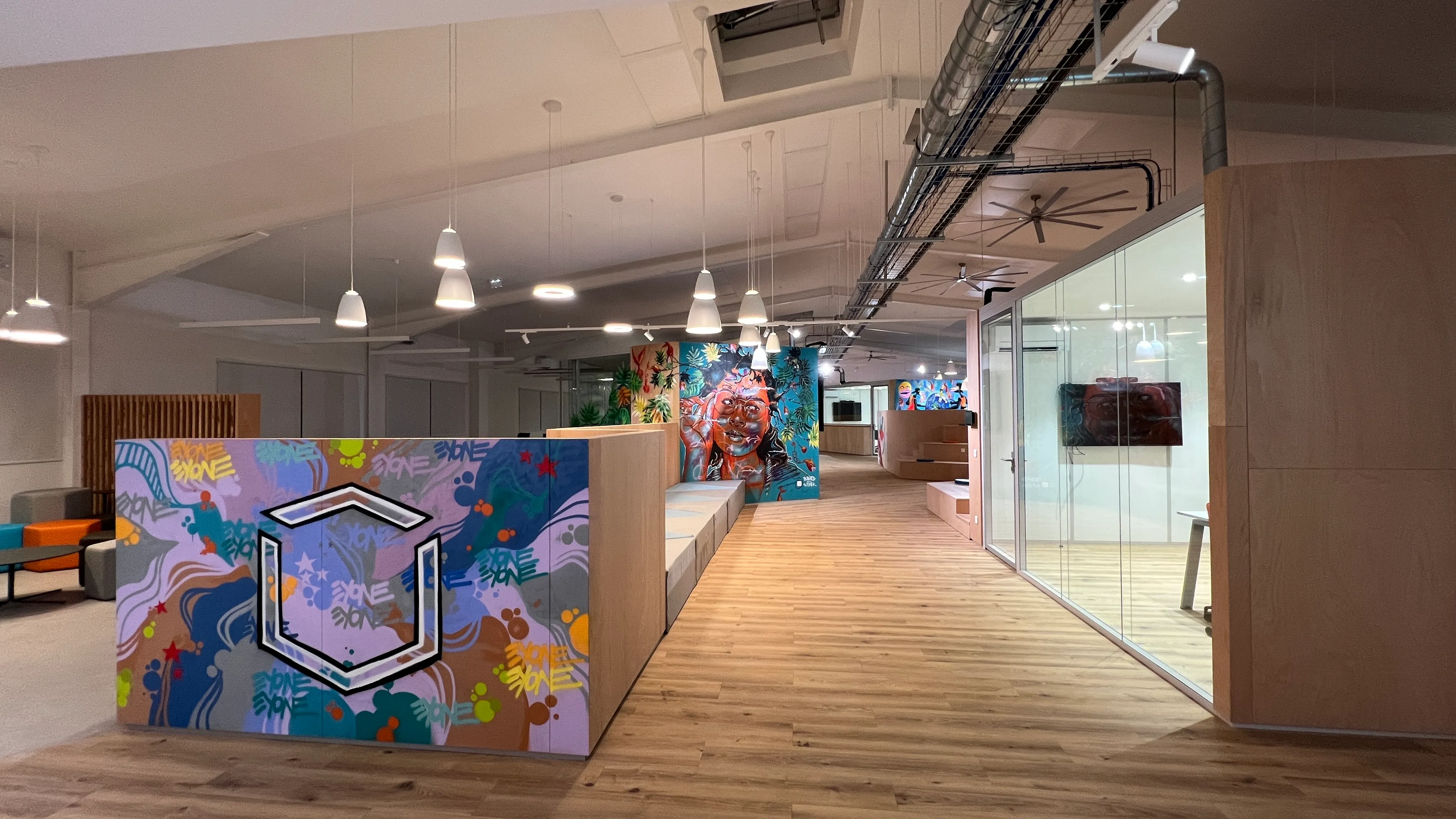 Interior of Zebox Guadeloupe with graffiti-style murals on partition walls, wood flooring and pendant lights leading toward glass-enclosed meeting cubicles