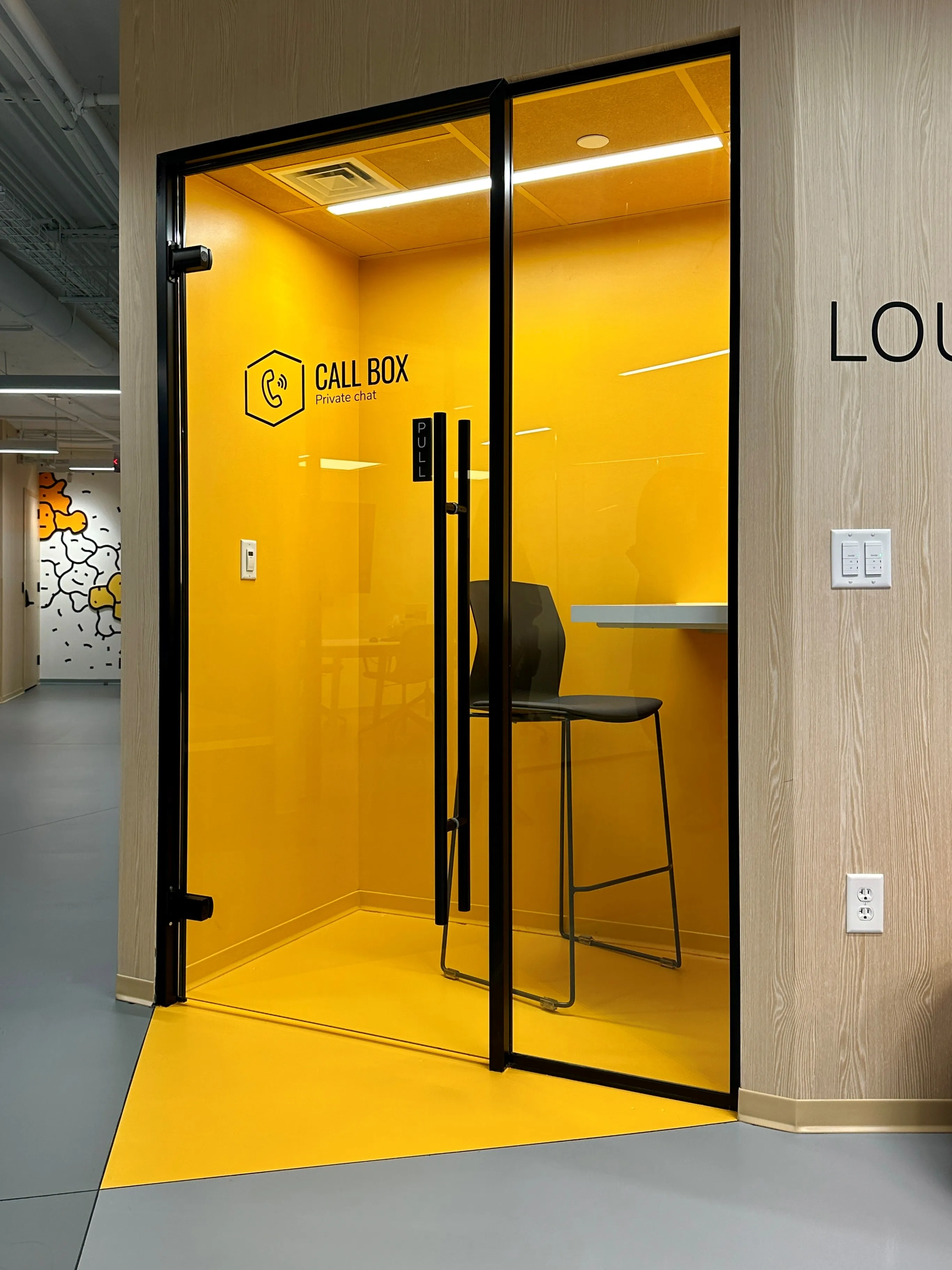 Glass-fronted phone booth with vivid yellow interior labeled "Call Box, Private chat" at Zebox Washington D.C., a stool and shelf inside