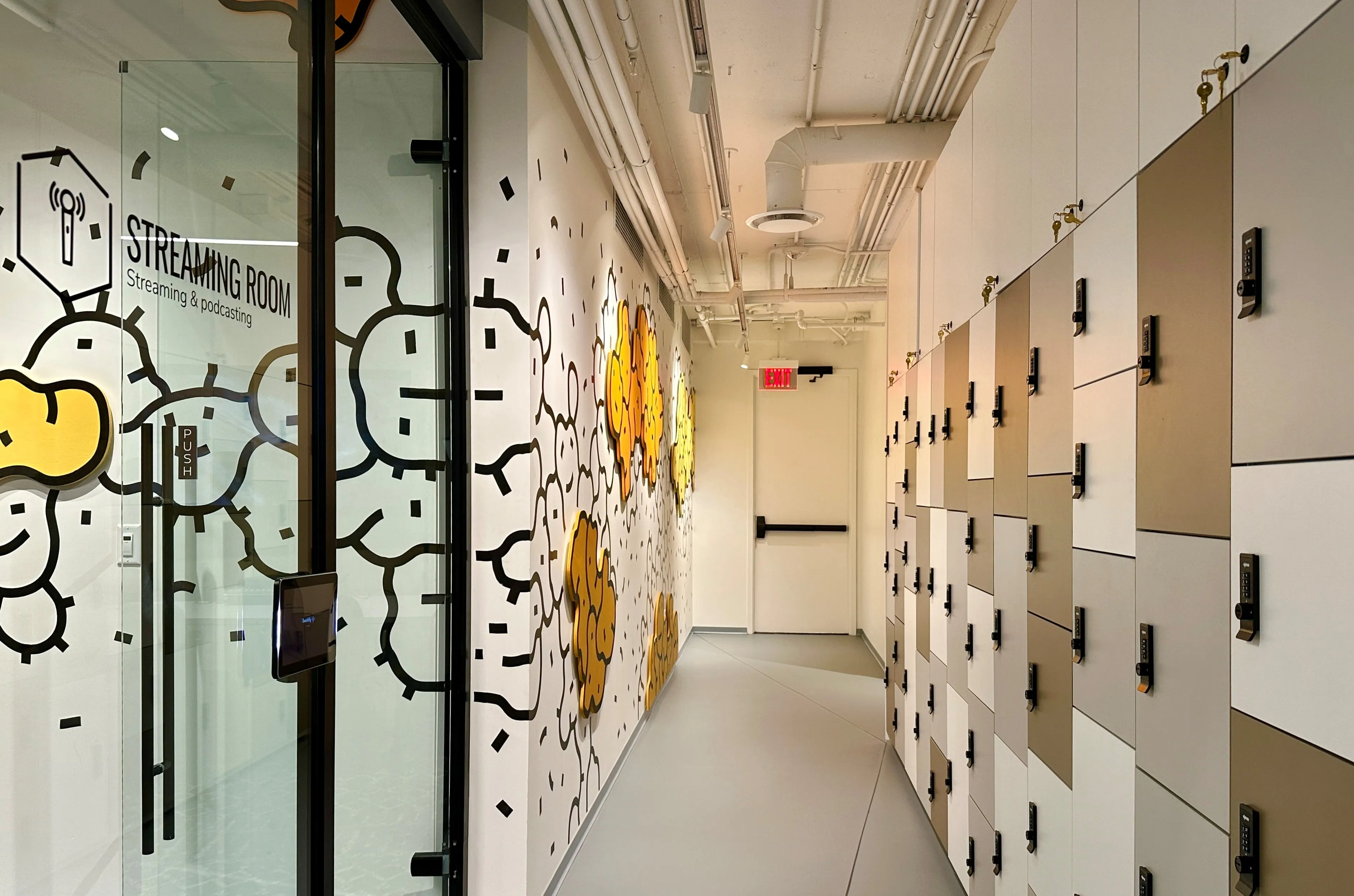 Corridor in Zebox Washington with a glass-walled streaming room, cloud-patterned mural, and a row of gold and white locker units along the hallway