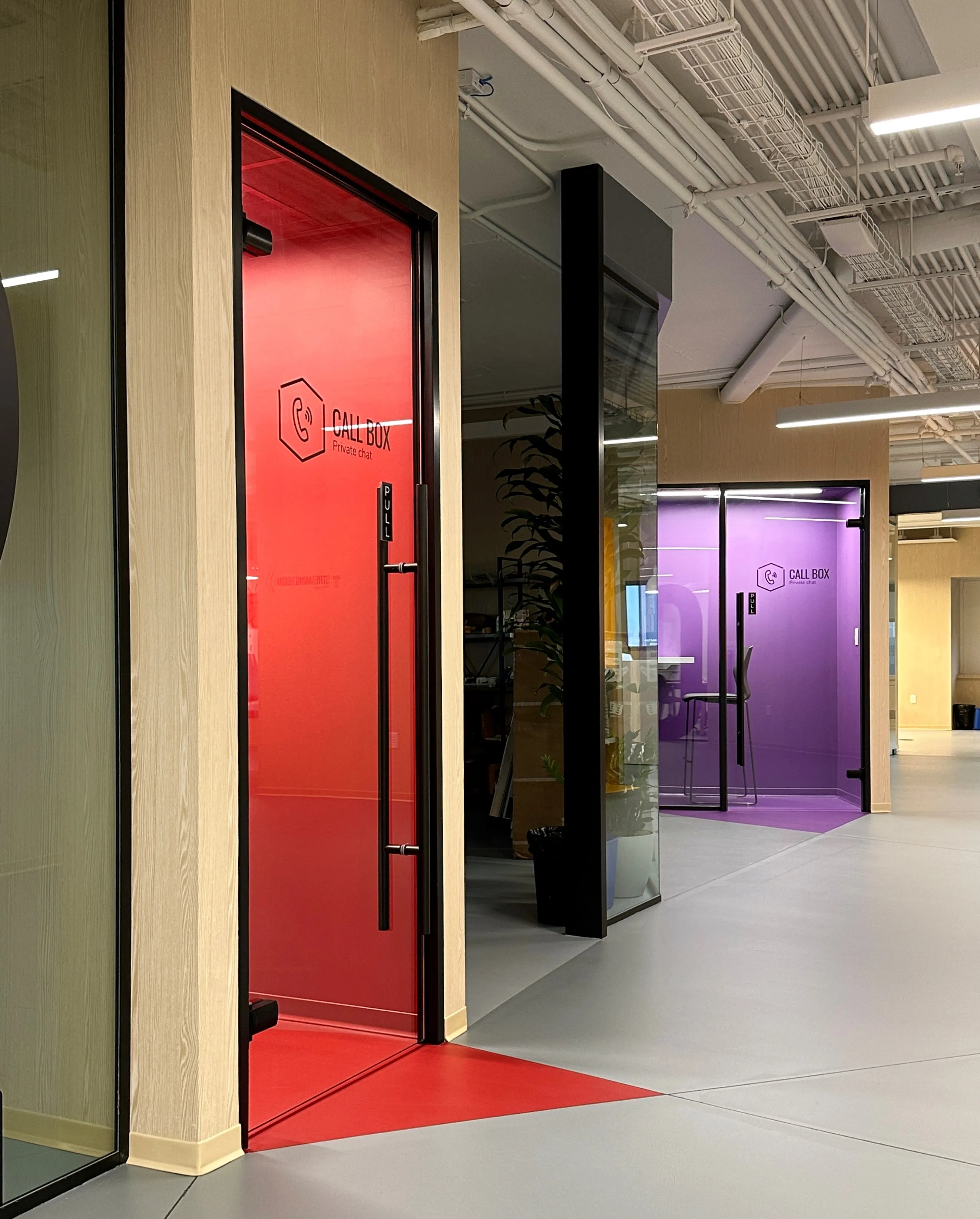 Red and purple glass-walled "Call Box" private phone booths line a corridor at Zebox Washington in Washington D.C., set beneath exposed ceiling ductwork