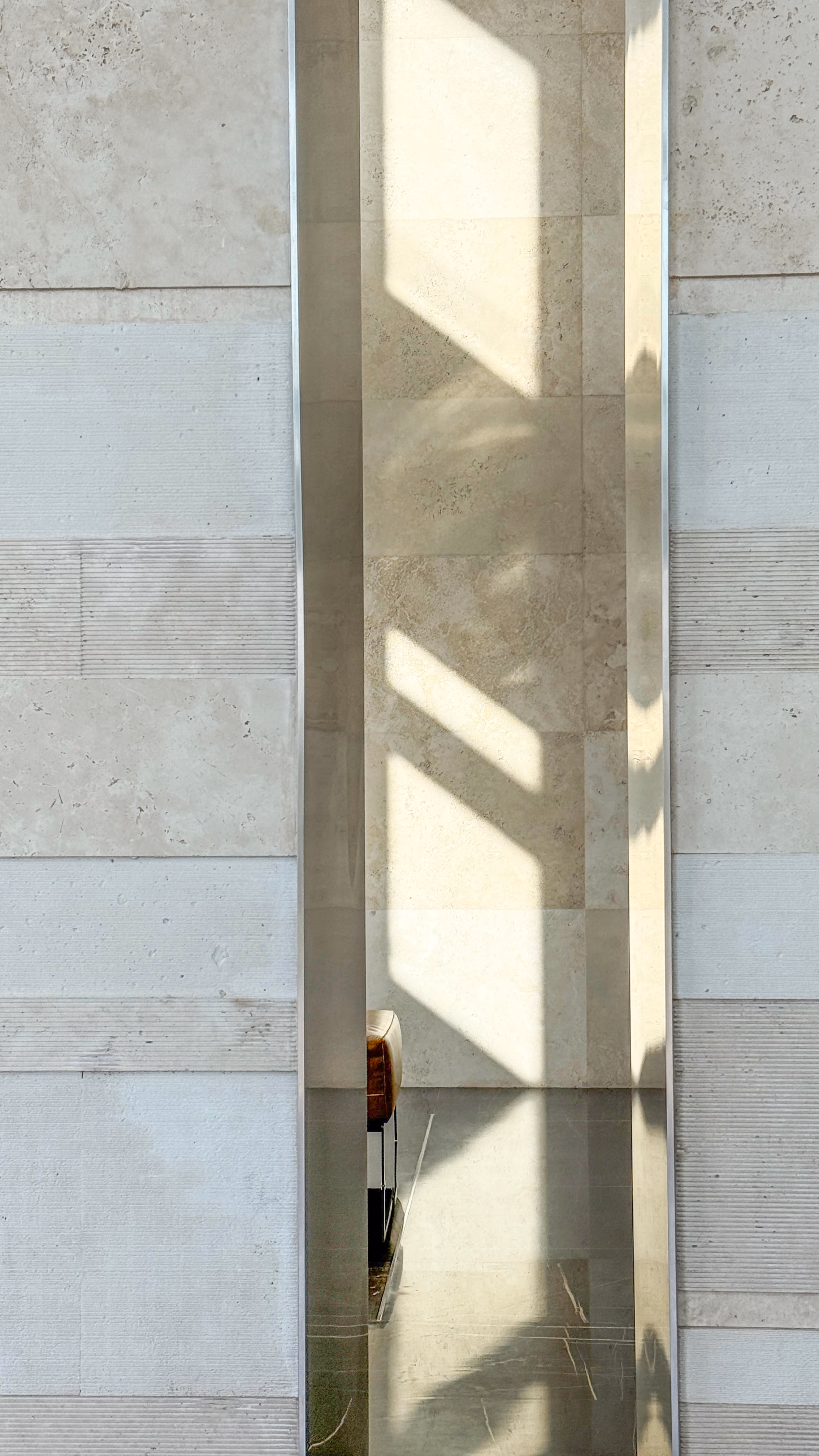 Narrow mirrored column in Golden Tower's Jeddah lobby, reflecting angled sunlight and travertine walls with subtly varied textured finishes