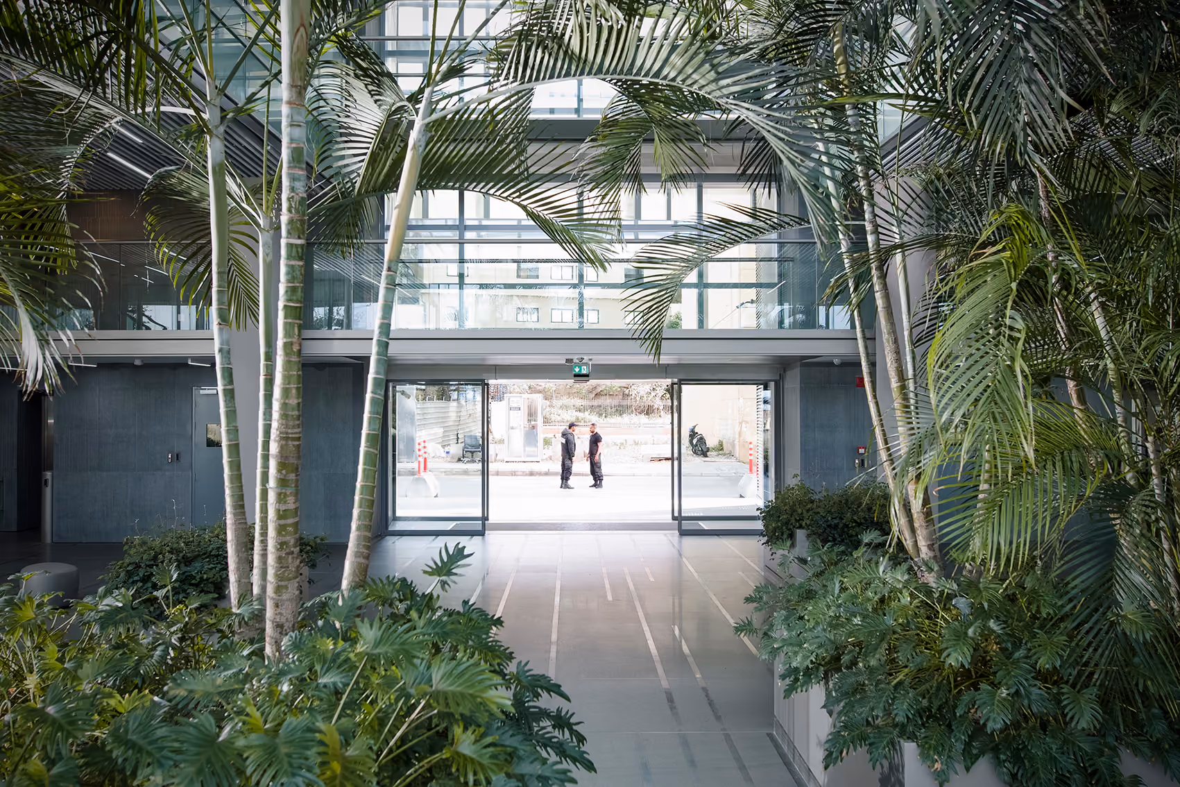 Palm-filled atrium of the CMA-CGM Headquarters refurbishment in Beirut, glazed bridges overhead framing a glass entrance where two people talk outside
