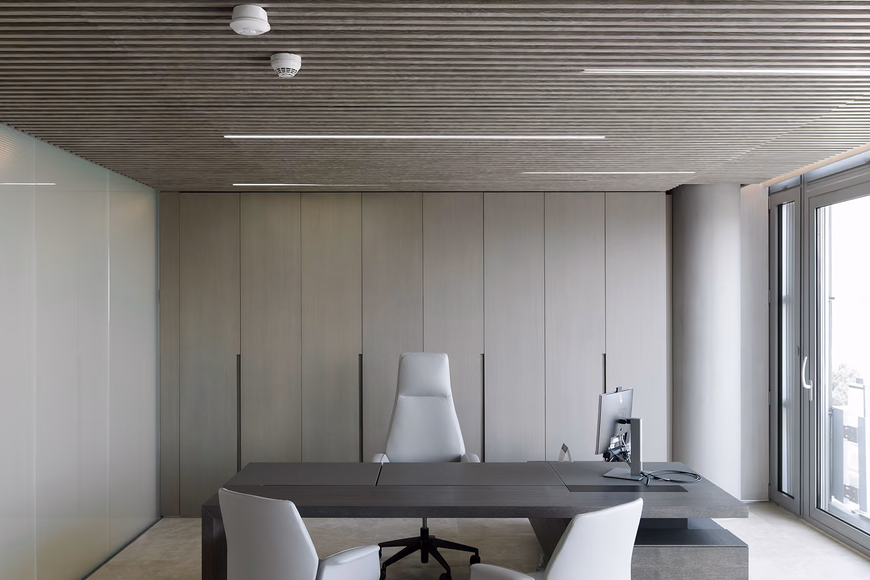 Executive office at CMA-CGM Headquarters in Beirut with ribbed wood-slat ceiling, grey panelled cabinetry, dark desk and white leather chairs beside a glazed balcony door