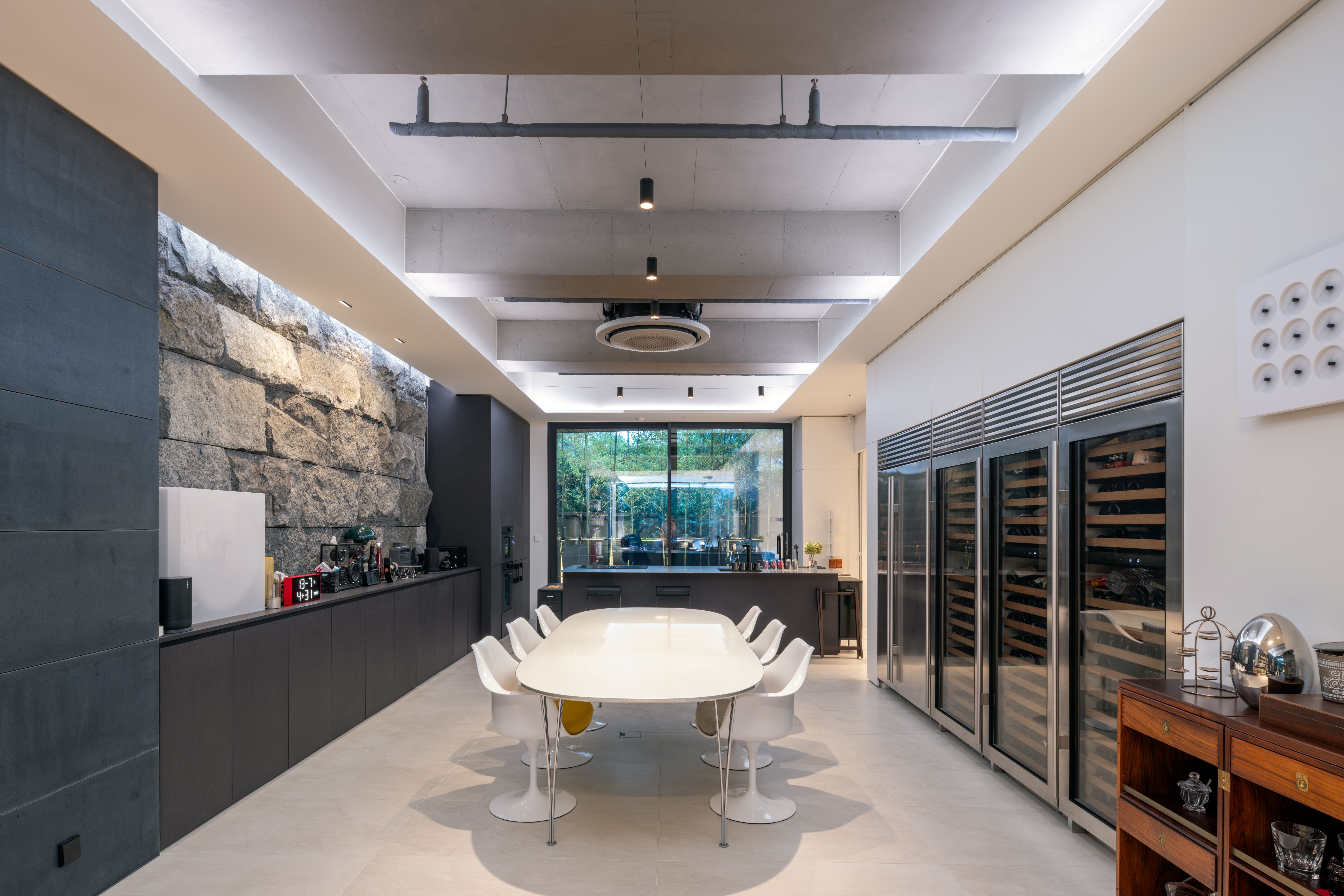 Dining and kitchen area in Pyrite house in Seoul, oval white table with tulip chairs beside a raw stone wall and built-in wine refrigerators