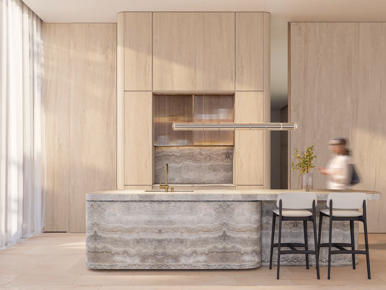 Kitchen in Nautile villa in Dubai, veined stone island with brass tap beneath light wood cabinetry and a ribbed glass niche, bar stools and a figure walking past