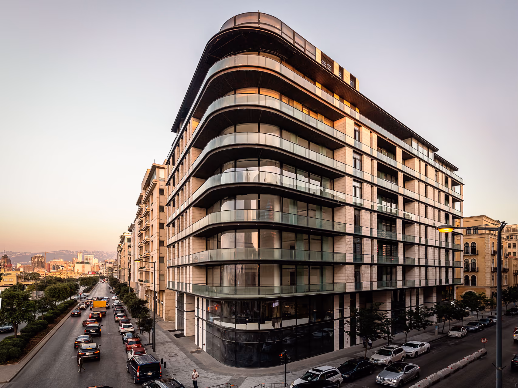 Allenby Gate in Beirut, corner view of a curved stone and glass residential building with stacked balconies overlooking a busy street at dusk
