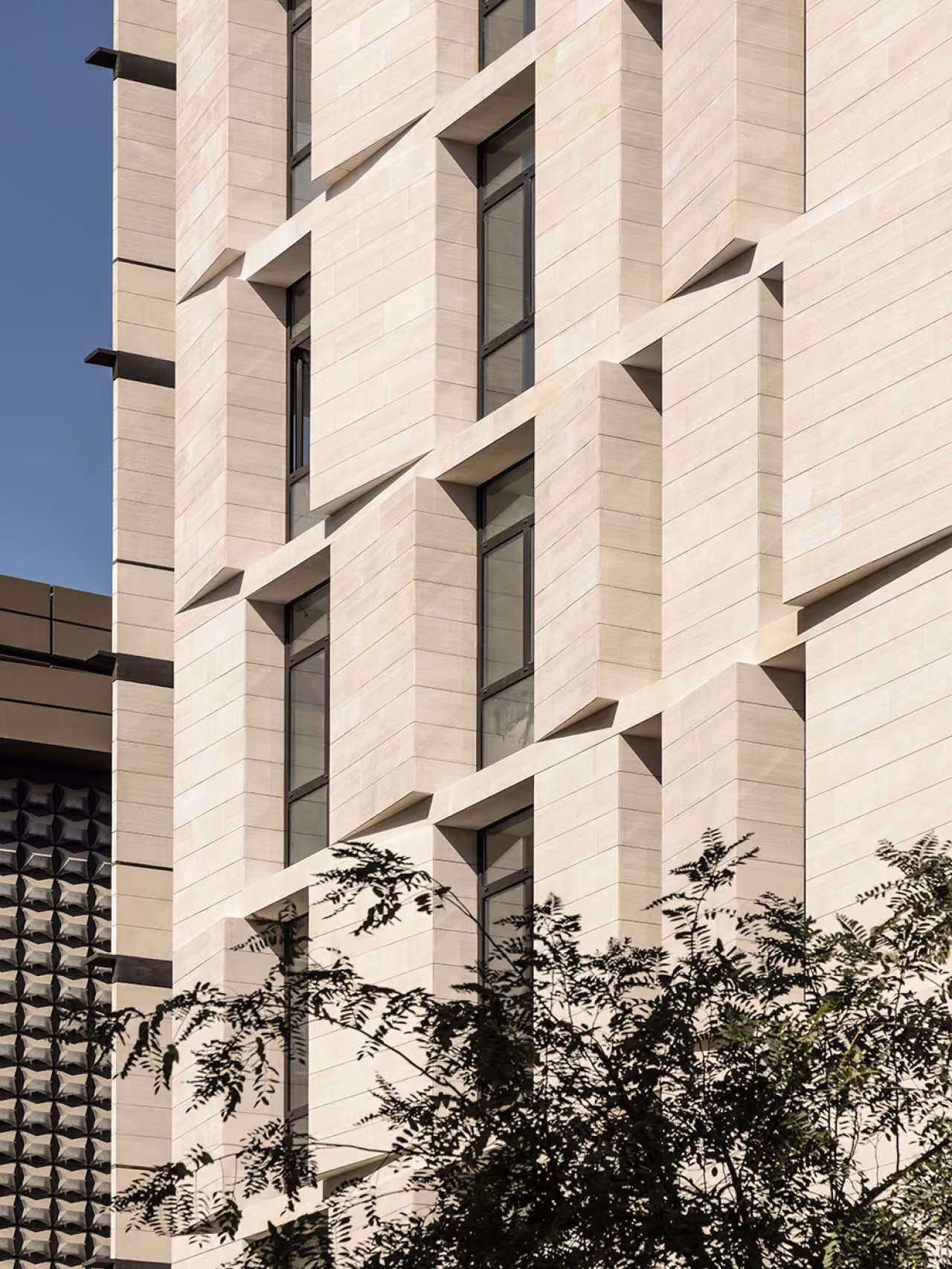 Facade detail of Allenby Gate in Beirut, angled stone-clad window bays creating a faceted pattern above trees