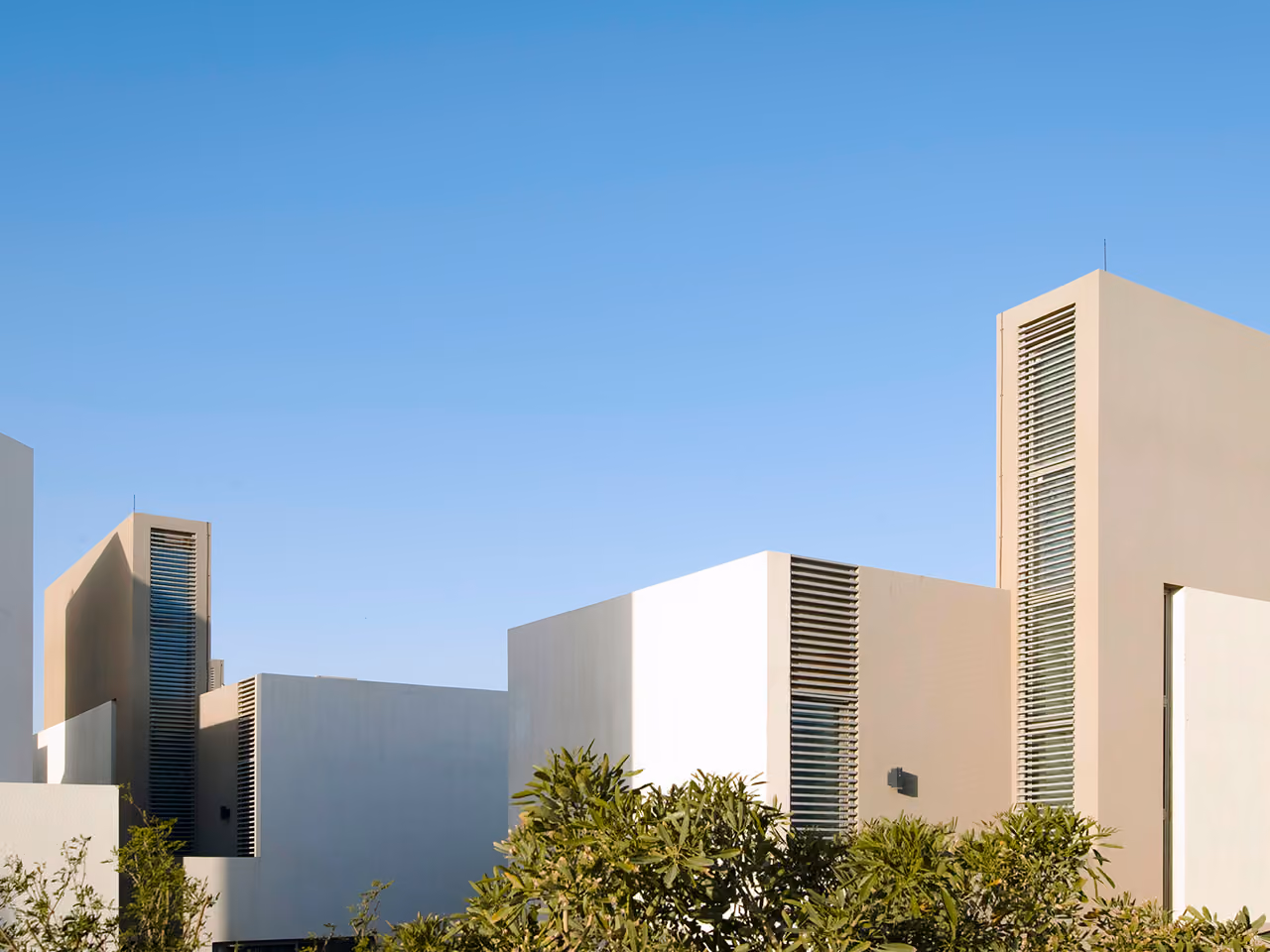 Cubic villas of Qortuba Oasis in Riyadh, staggered beige and white volumes with vertical louvred shafts against a clear sky and lush shrubs