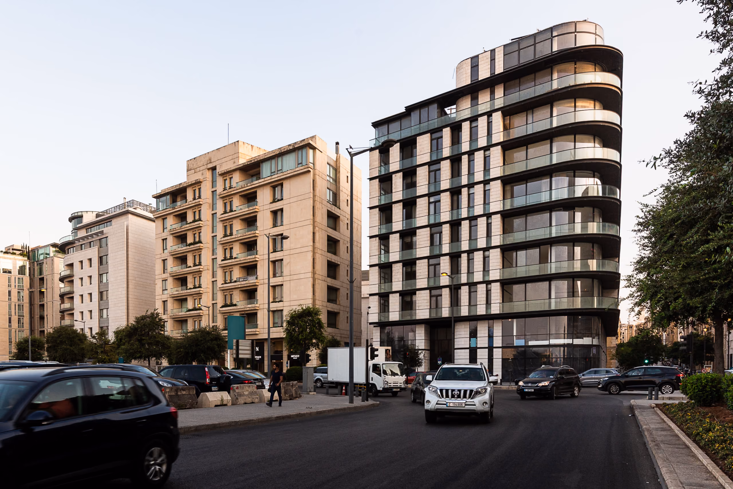 Allenby Gate in Beirut, a curved corner tower with rounded balconies and glass facade, standing among stone-clad midrise buildings on a busy street