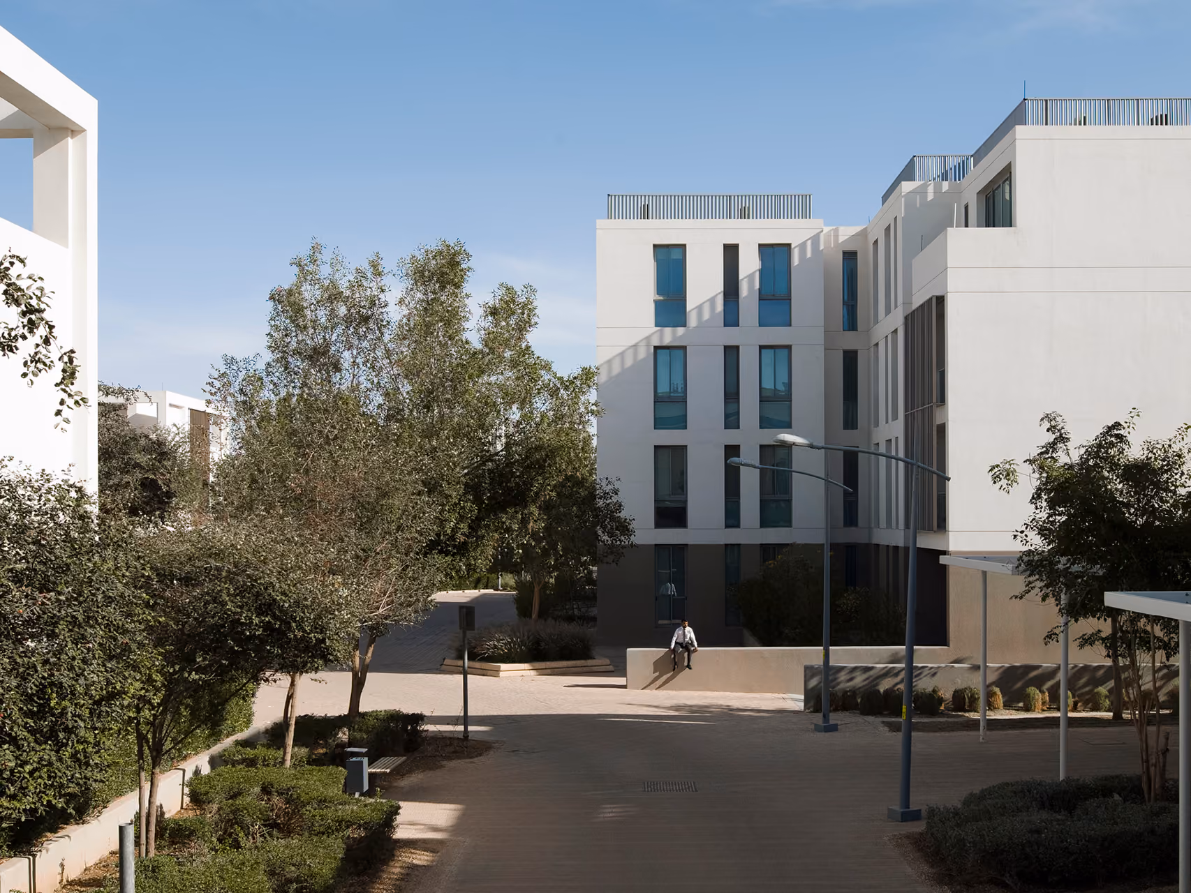 Tree-lined pathway at Qortuba Oasis in Riyadh, leading to a white low-rise apartment block with recessed balconies and a seated figure resting on a low wall