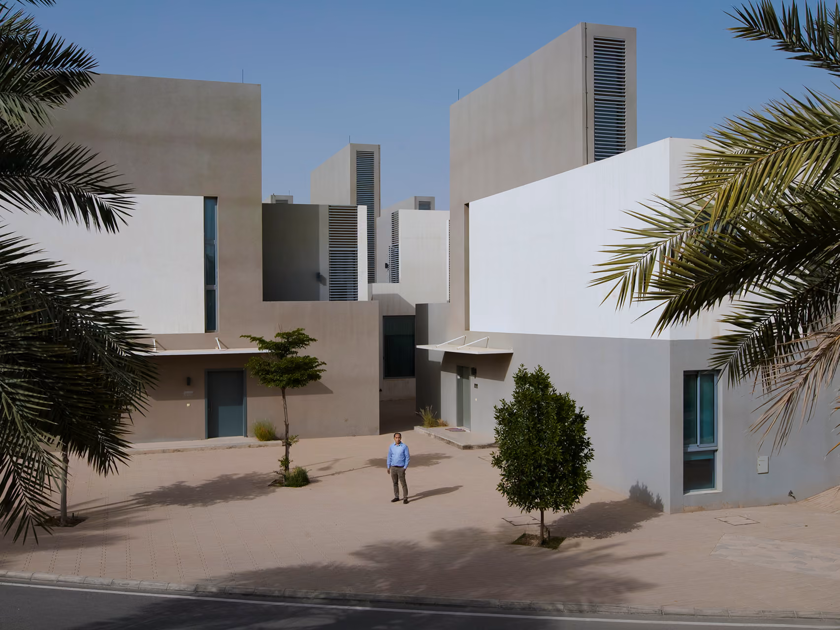 Cluster of angular concrete villas at Qortuba Oasis in Riyadh, tall ventilation towers rising above a courtyard with palm trees and a figure walking through