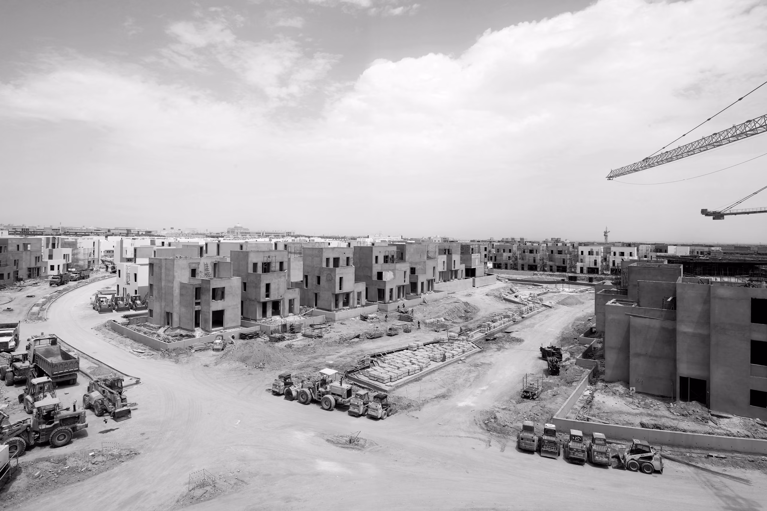 Black and white construction view of Qortuba Oasis in Riyadh, rows of unfinished concrete villas with cranes, bulldozers and graders across the site