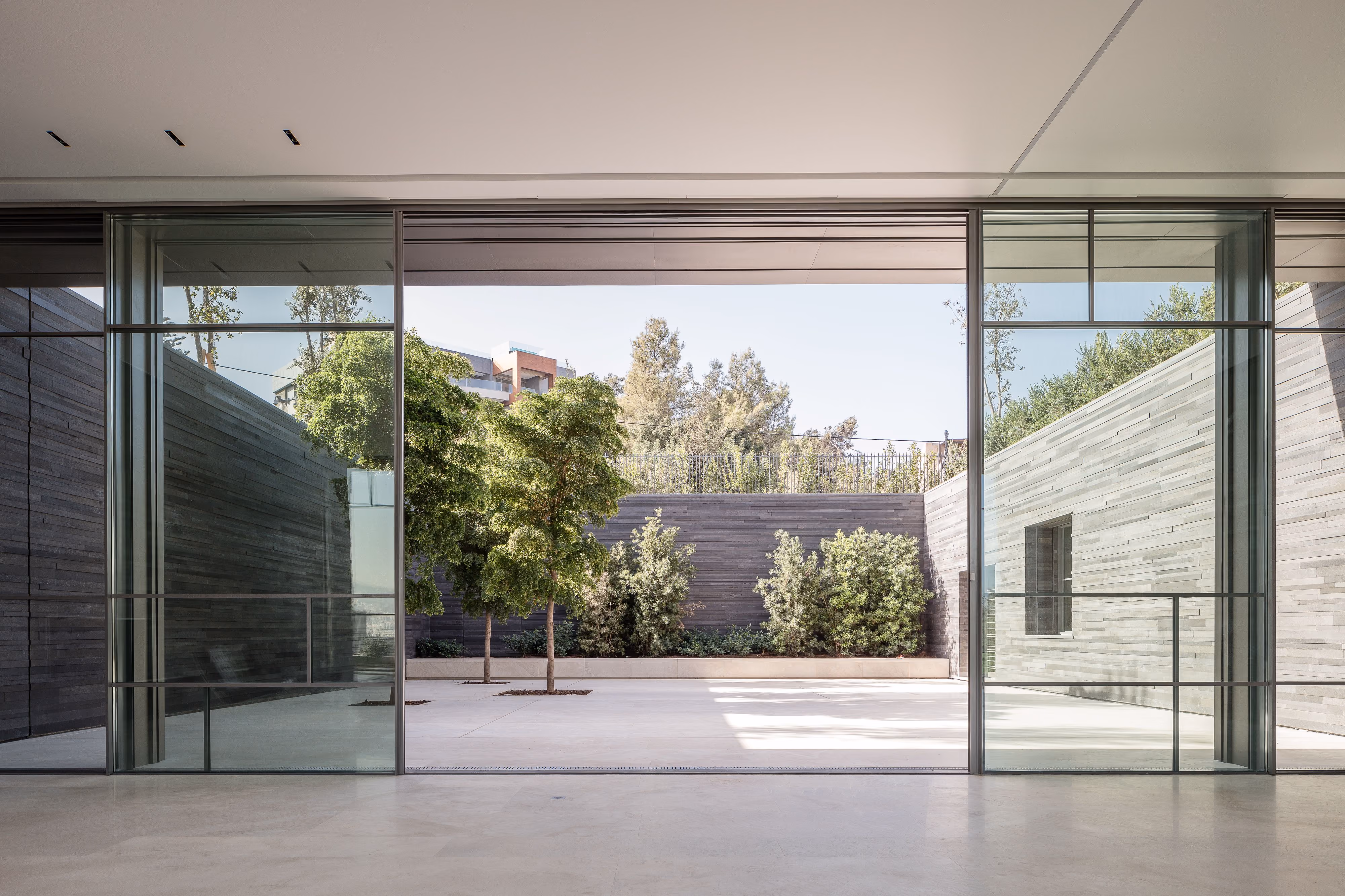 Glass-walled opening in AY House in Yarze framing a stone-paved courtyard with slender trees against dark basalt walls