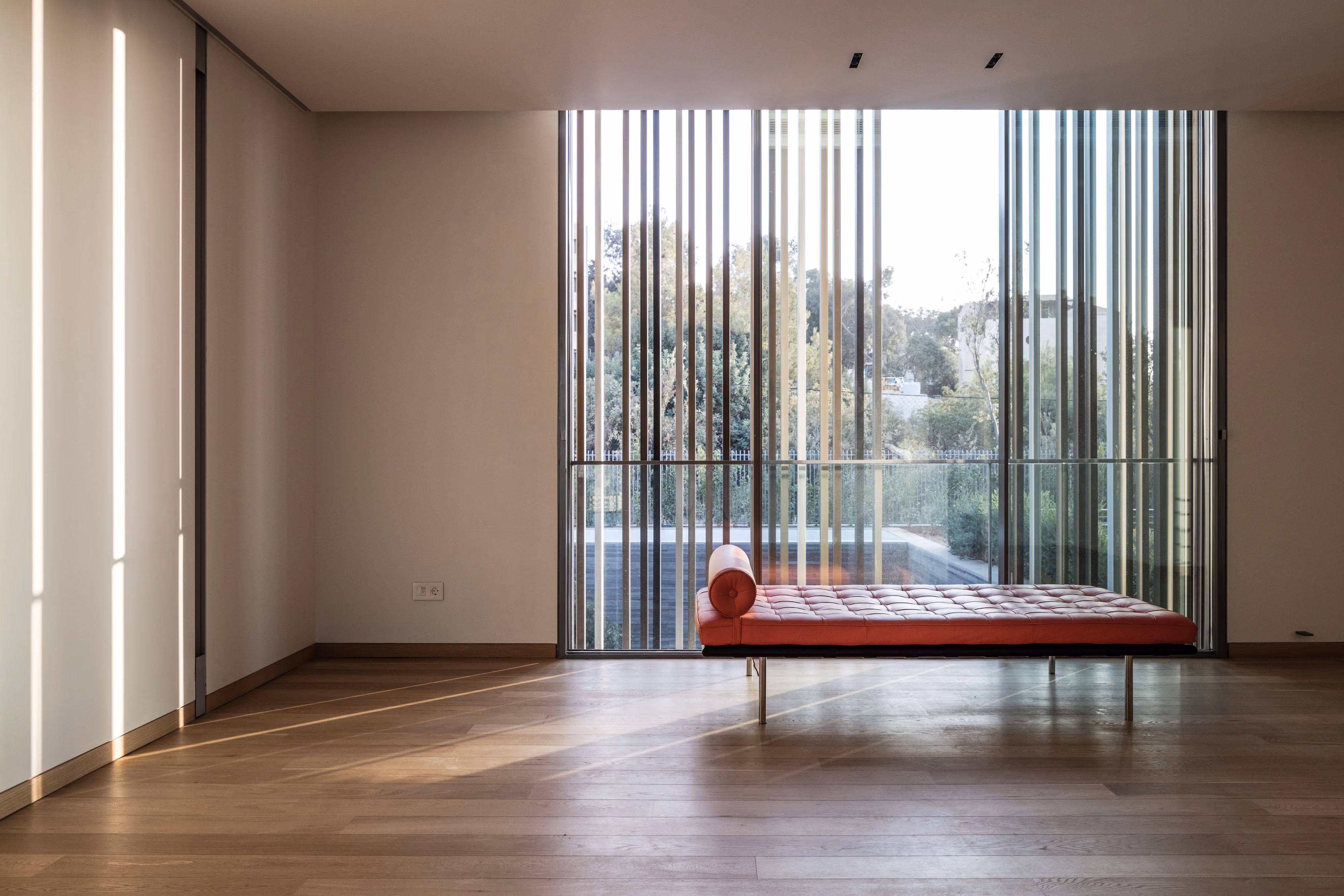 Interior of AY House in Yarze, red tufted daybed by a floor-to-ceiling louvered glass wall casting light patterns on timber flooring