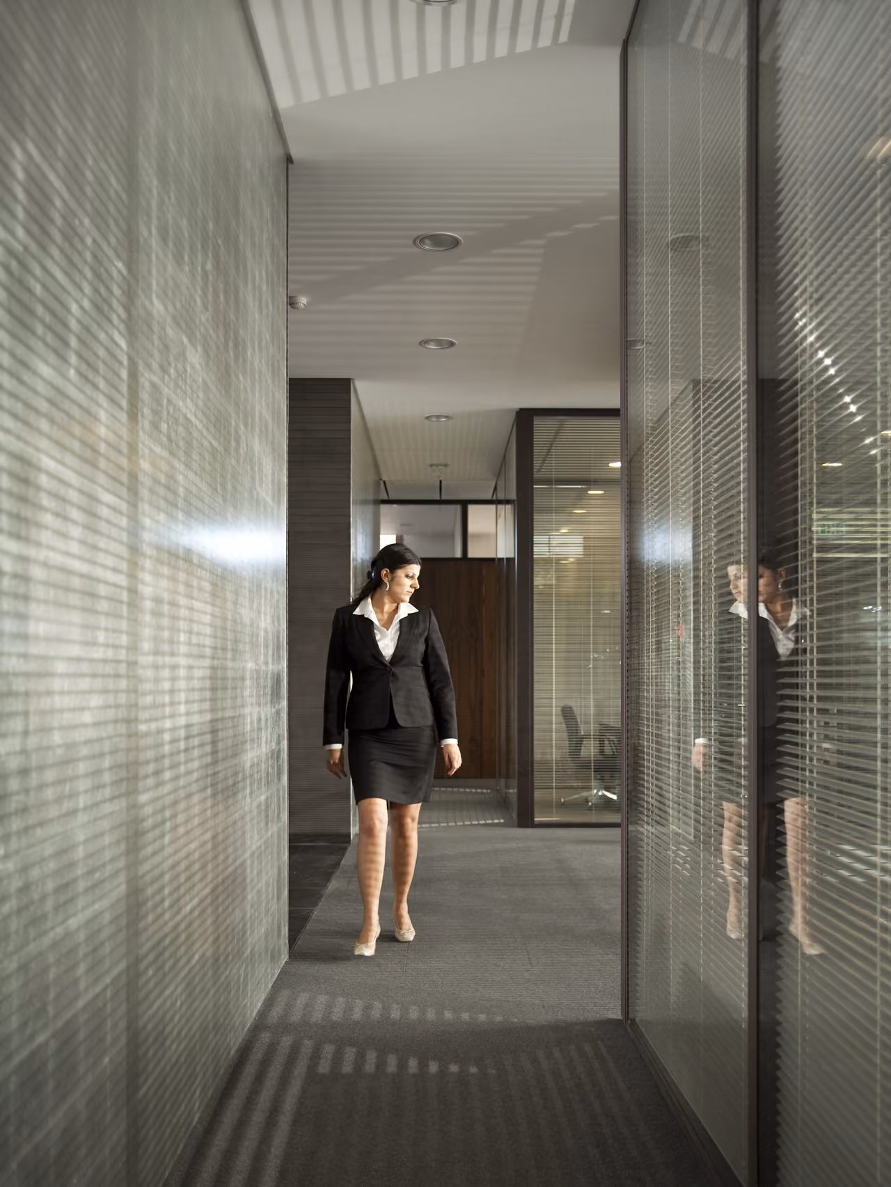 A woman in a dark suit walks a glass-lined corridor at Waqf Foch Office Building in Beirut, textured walls reflecting striped light patterns