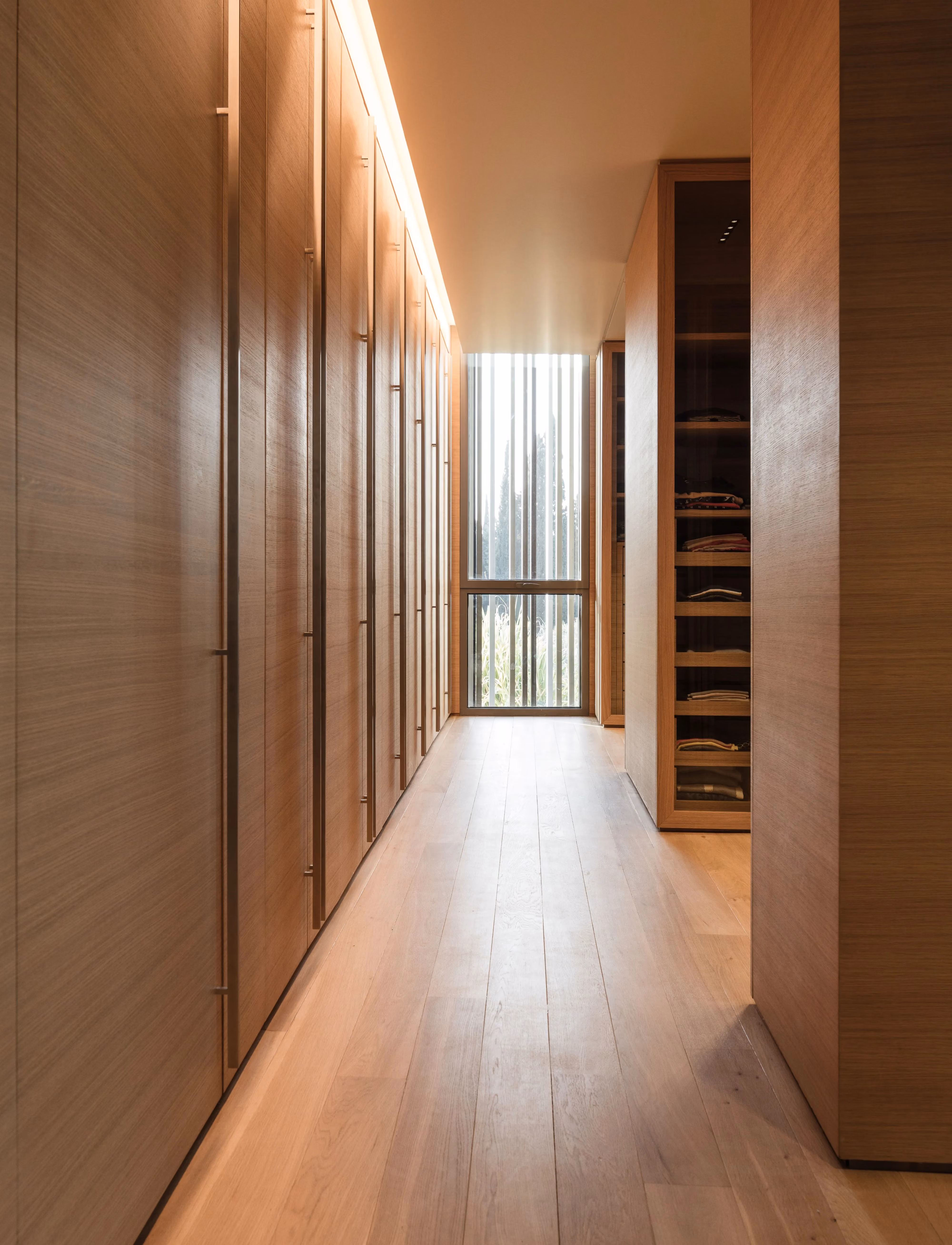 Walk-in closet at AY House in Yarze, wood-panelled wardrobes lining a corridor toward a louvred window with timber flooring