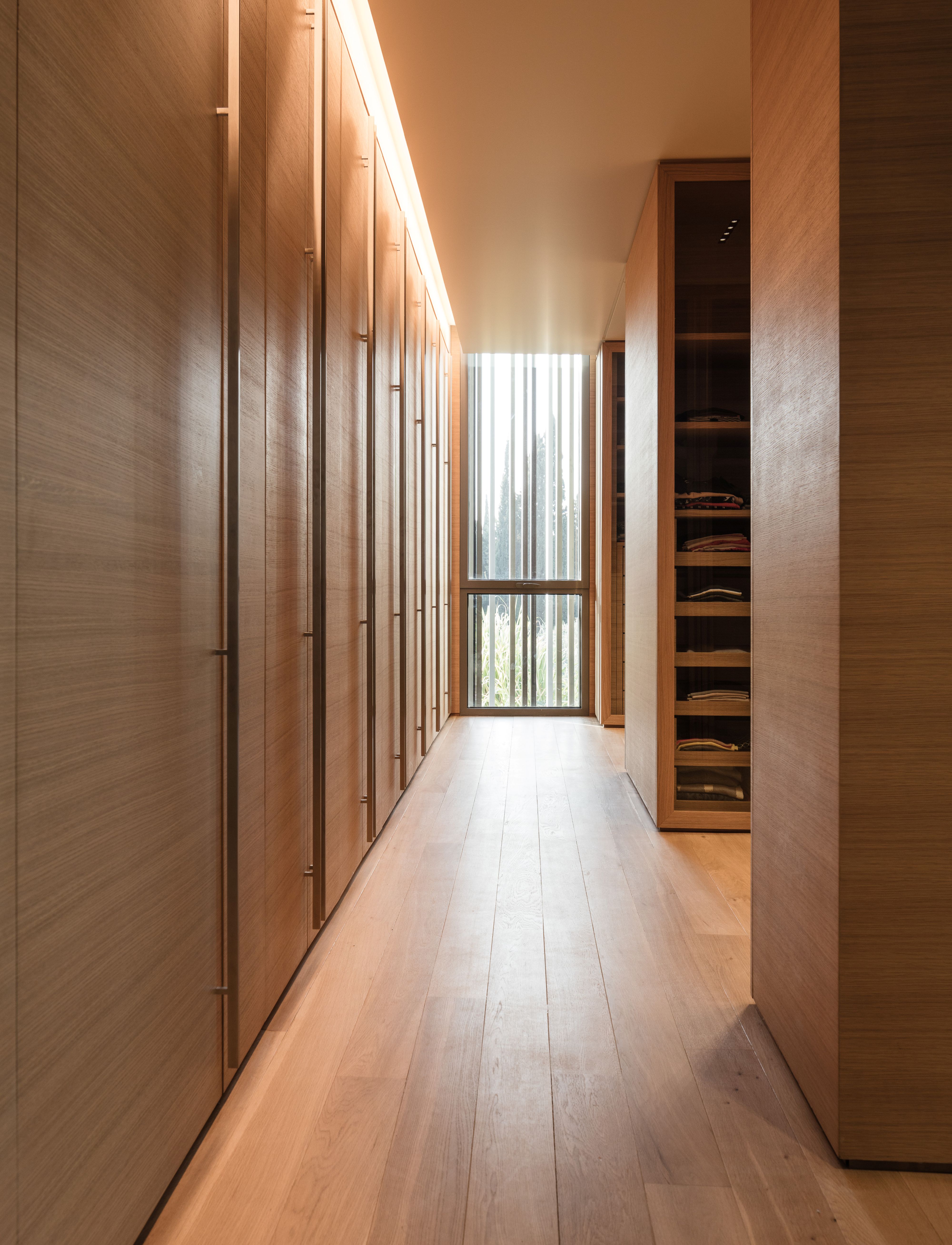 Walk-in closet at AY House in Yarze, wood-panelled wardrobes lining a corridor toward a louvred window with timber flooring