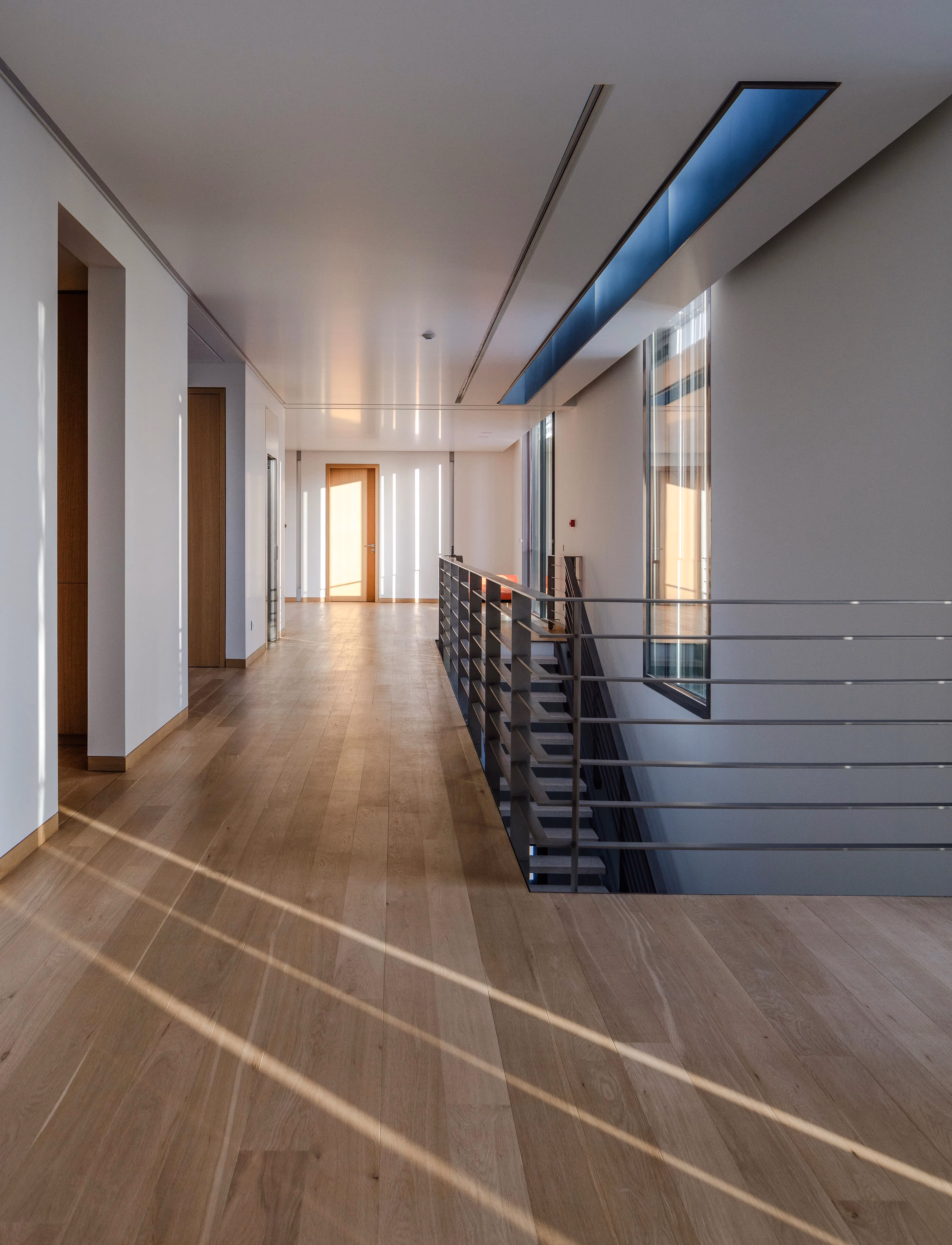 Upper-level corridor at AY House in Yarze, oak flooring with sunlit stripes, open stairwell railing and a skylight casting a blue-tinted beam along the ceiling