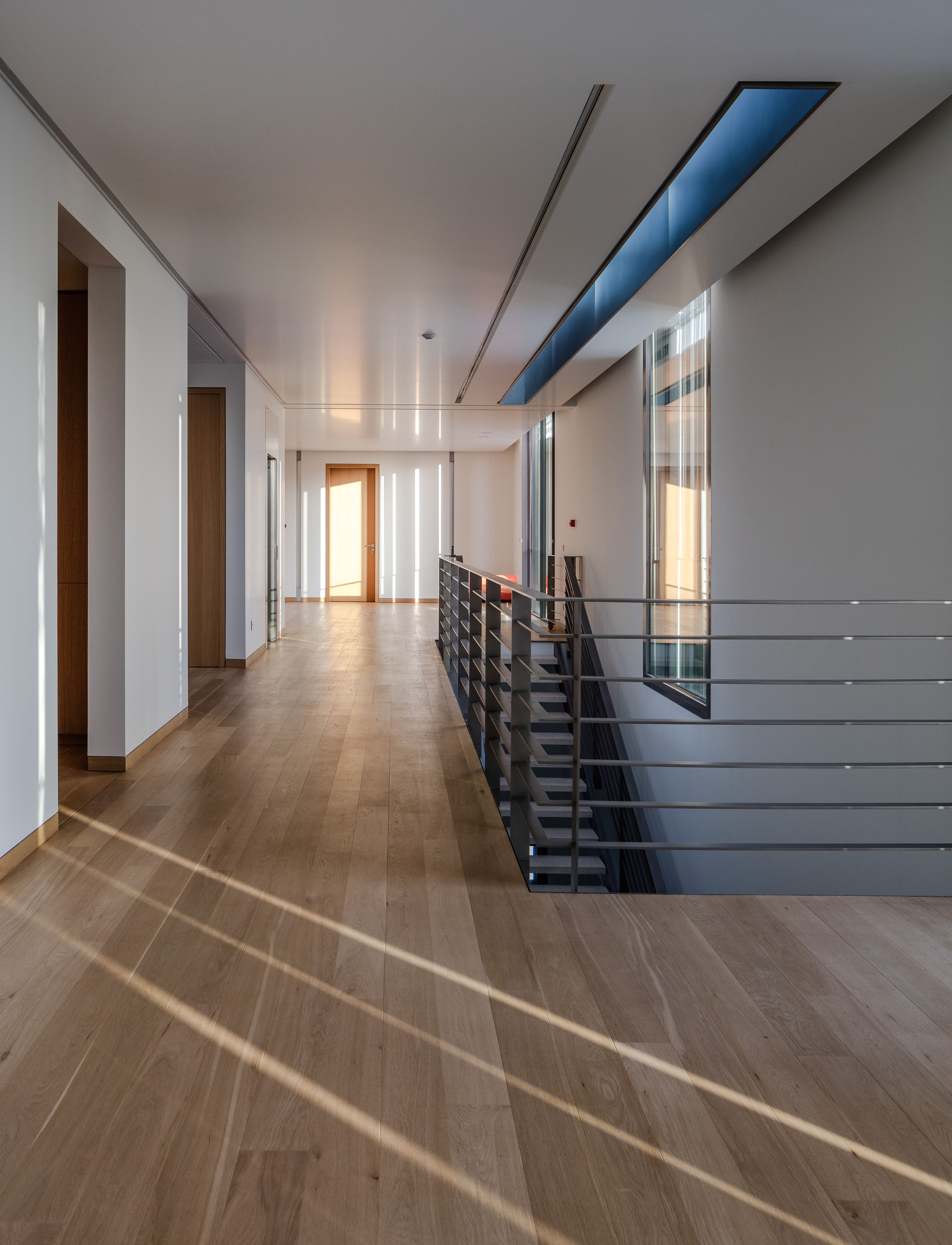 Upper-level corridor at AY House in Yarze, oak flooring with sunlit stripes, open stairwell railing and a skylight casting a blue-tinted beam along the ceiling