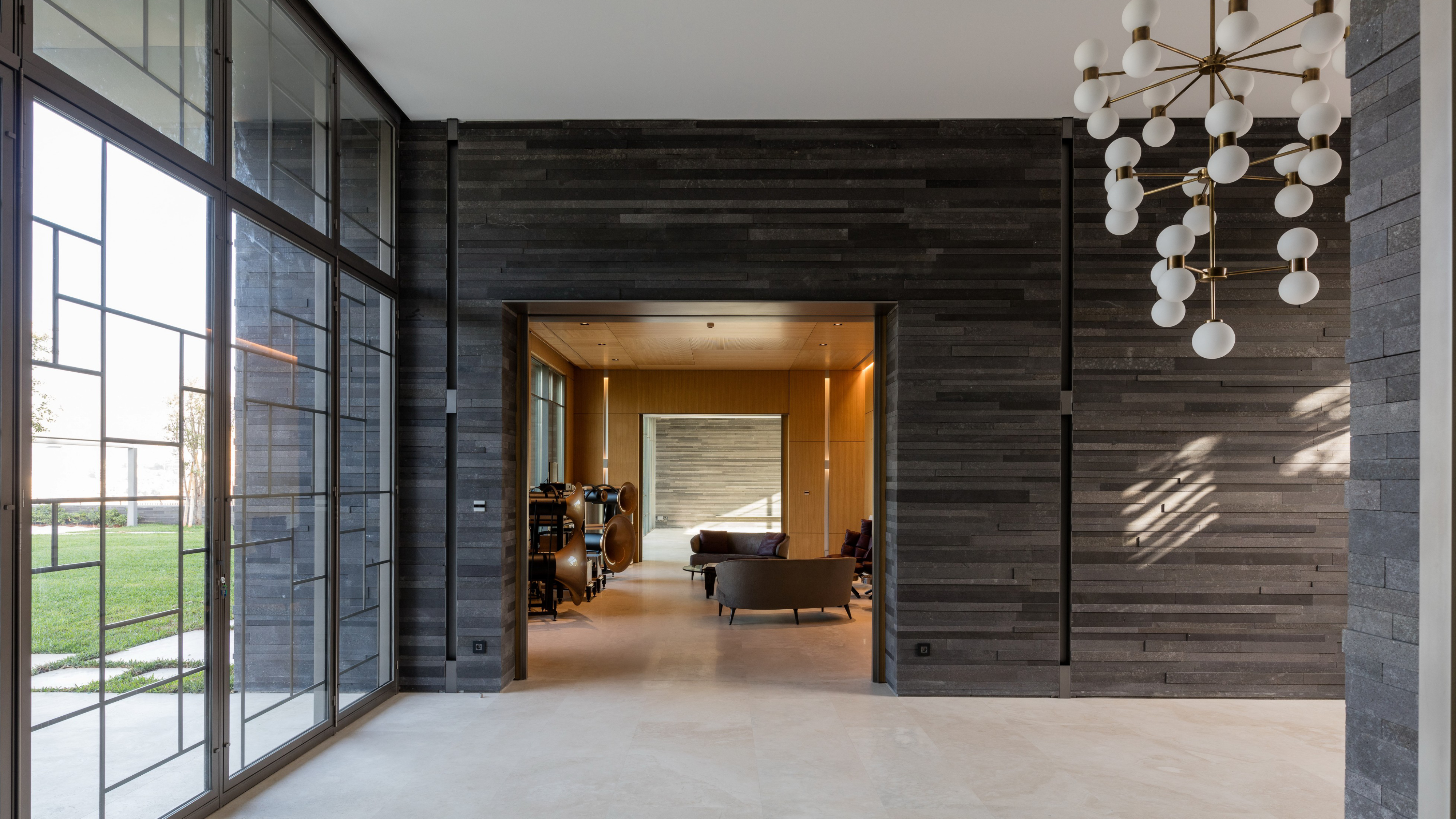 AY House interior design in Yarze, Lebanon, by Nabil Gholam Architects, layered basalt-clad walls, floor-to-ceiling glazing and a sculptural globe chandelier leading to a warm lounge beyond