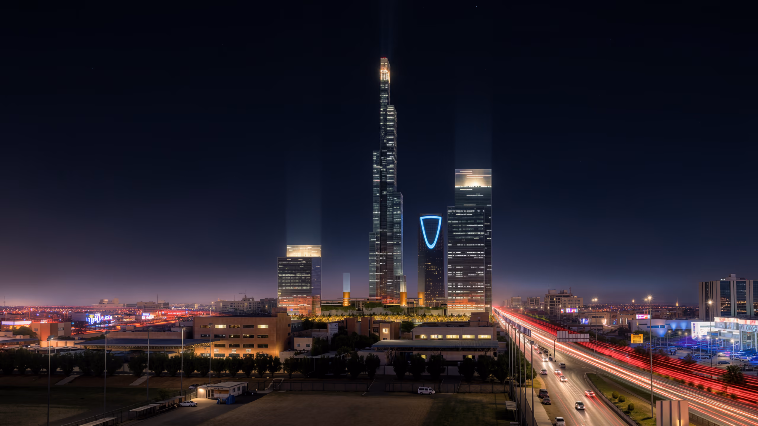 Night view of the Mixed-Use Development in Riyadh, its slender illuminated tower rising above the skyline beside the Kingdom Centre with light trails from traffic below