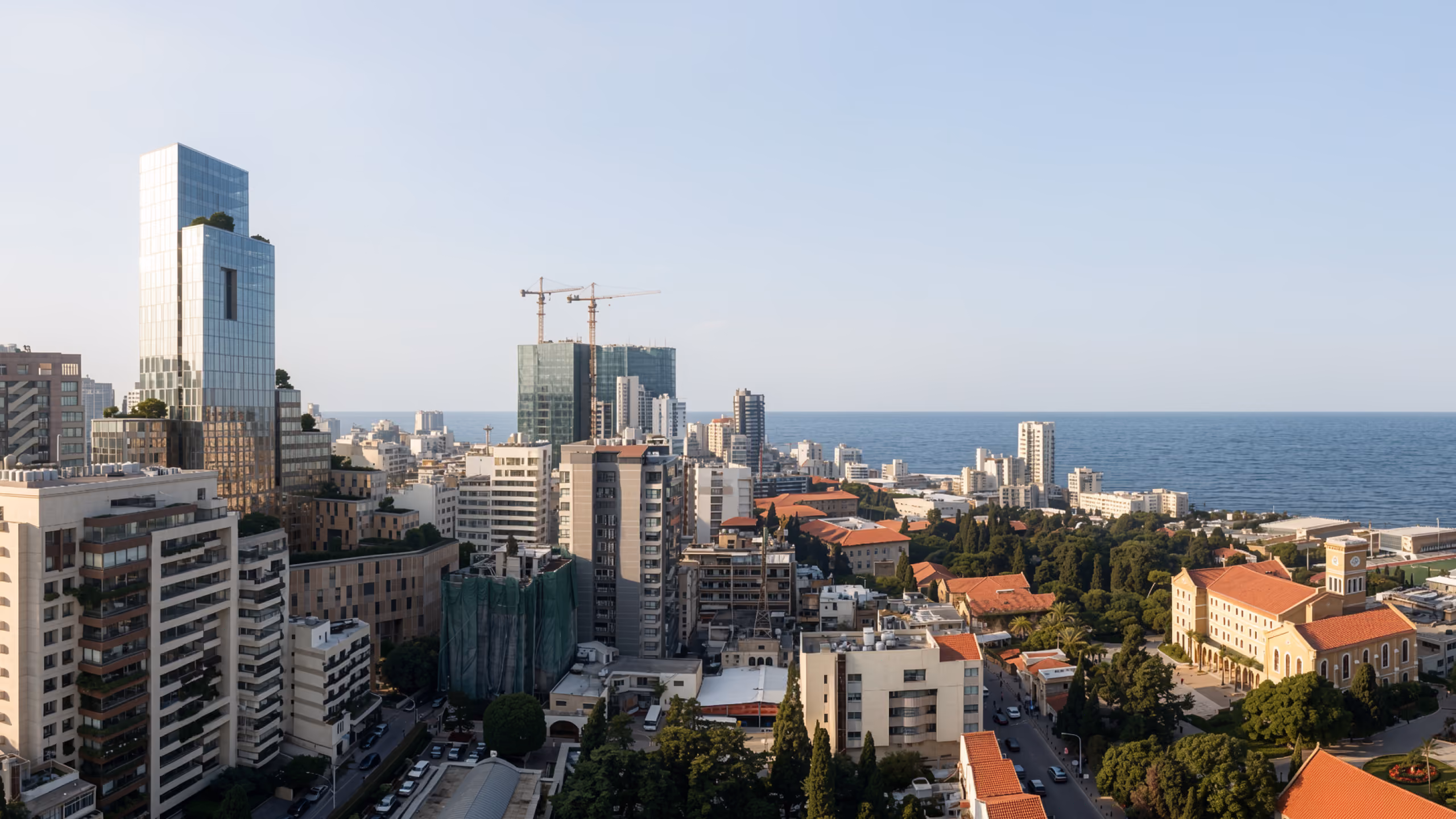 City Campus 01 in Beirut, aerial view of the dense coastal skyline with the mirrored glass tower rising above red-roofed low-rise buildings and the sea beyond