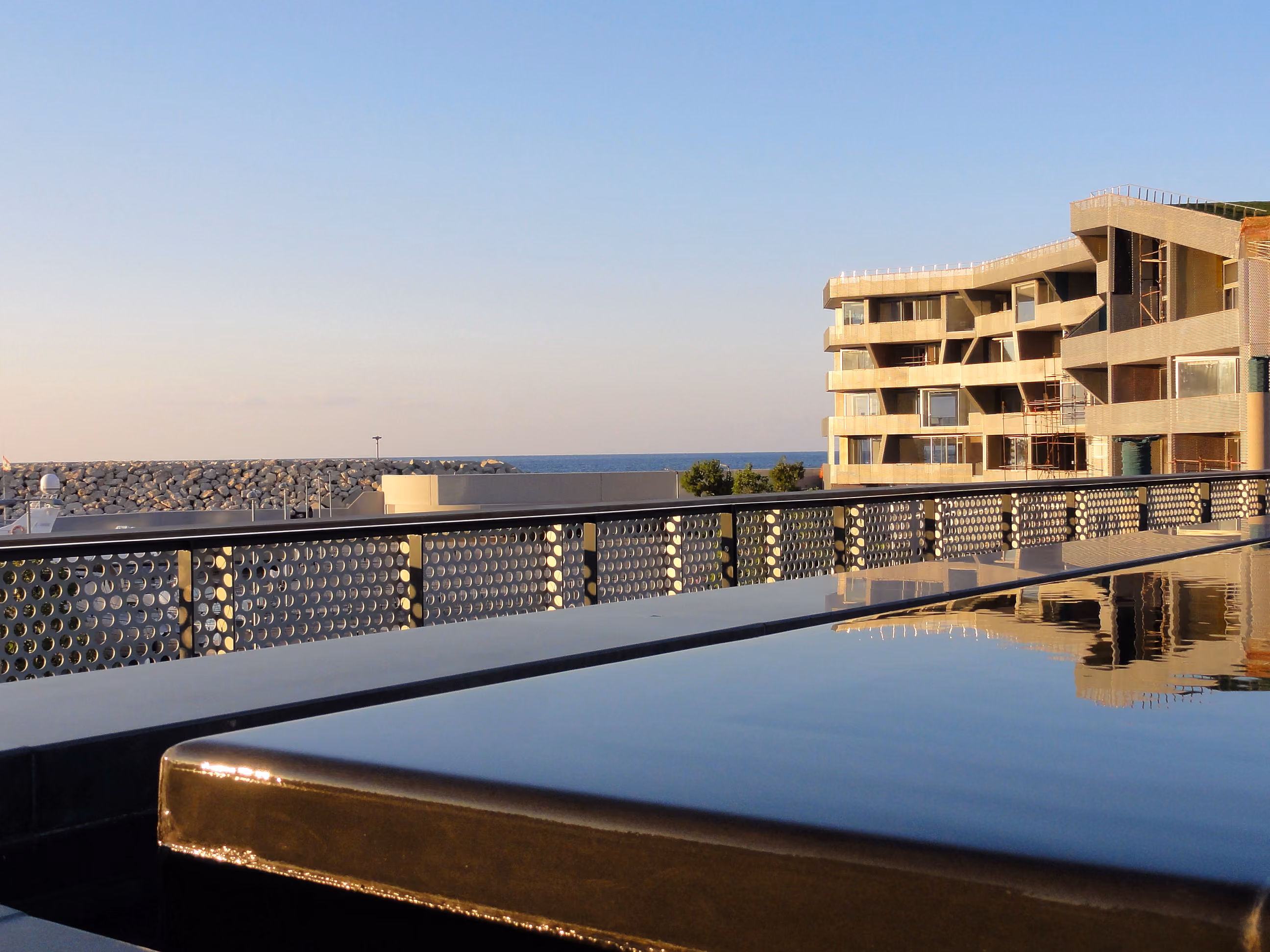 Rooftop pool with perforated metal railing at Beirut Marina & Town Quay, overlooking a breakwater, the sea and a terraced concrete building under construction