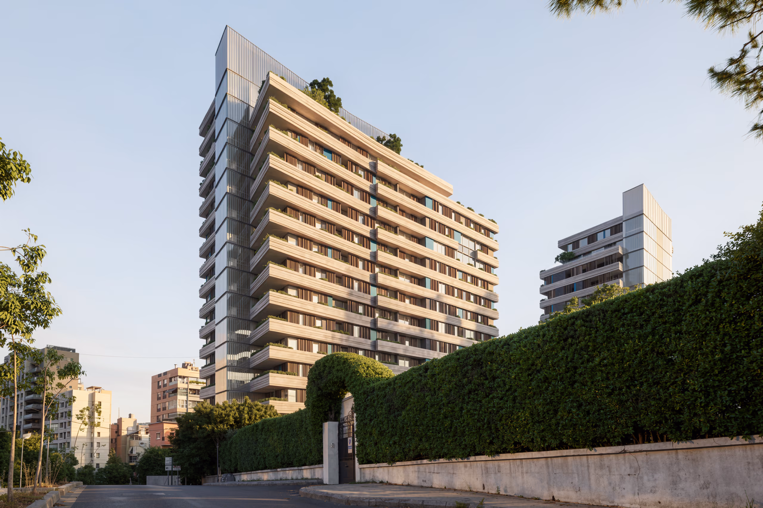 679, residential tower in Beirut's Raouché district, its stacked terraces and vertical stone louvres rising above a hedge-lined street and neighbouring buildings