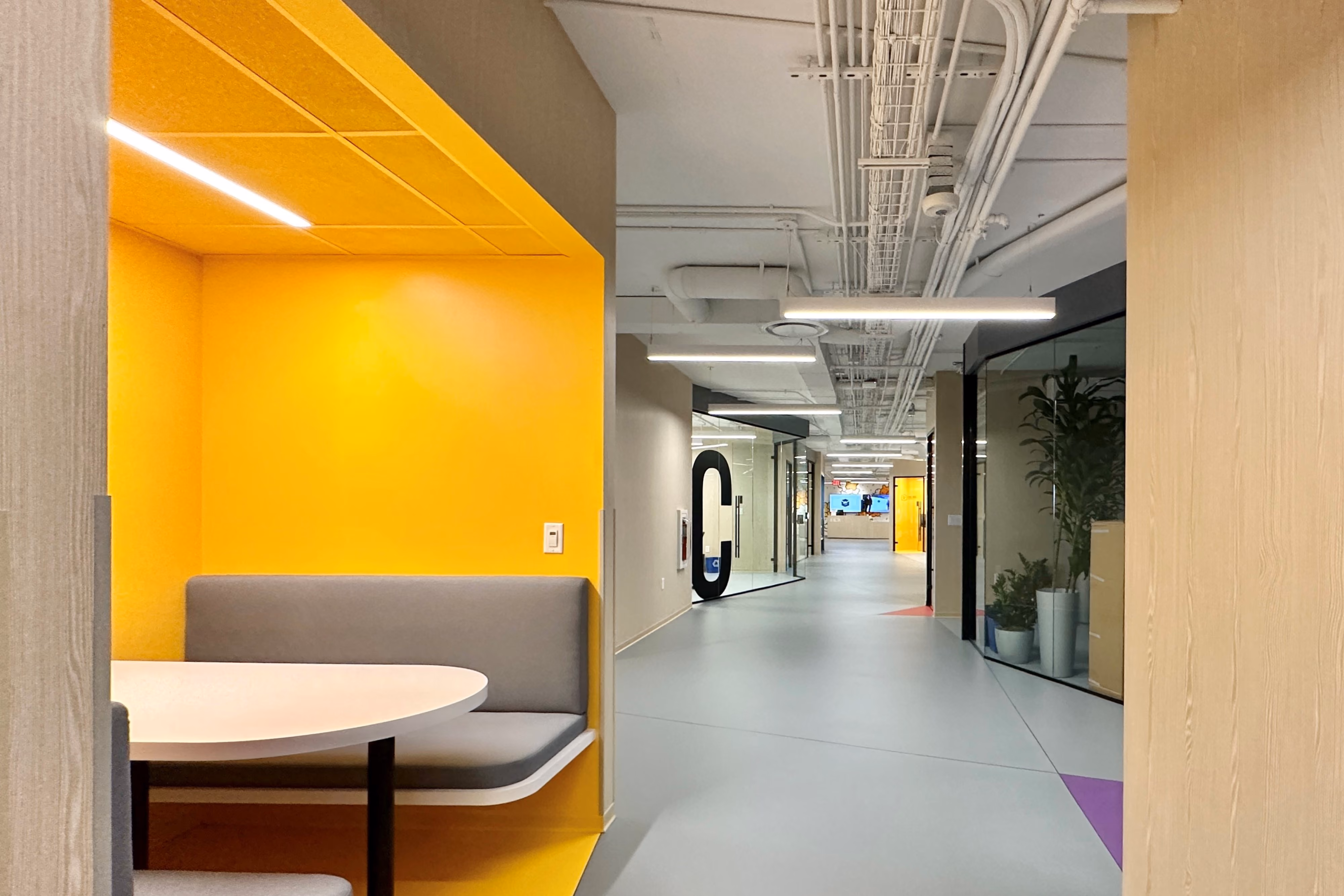 Zebox Washington, office interior by Nabil Gholam Architects, yellow booth nook opening onto corridor of exposed ceilings and glass-walled rooms in Washington D.C.