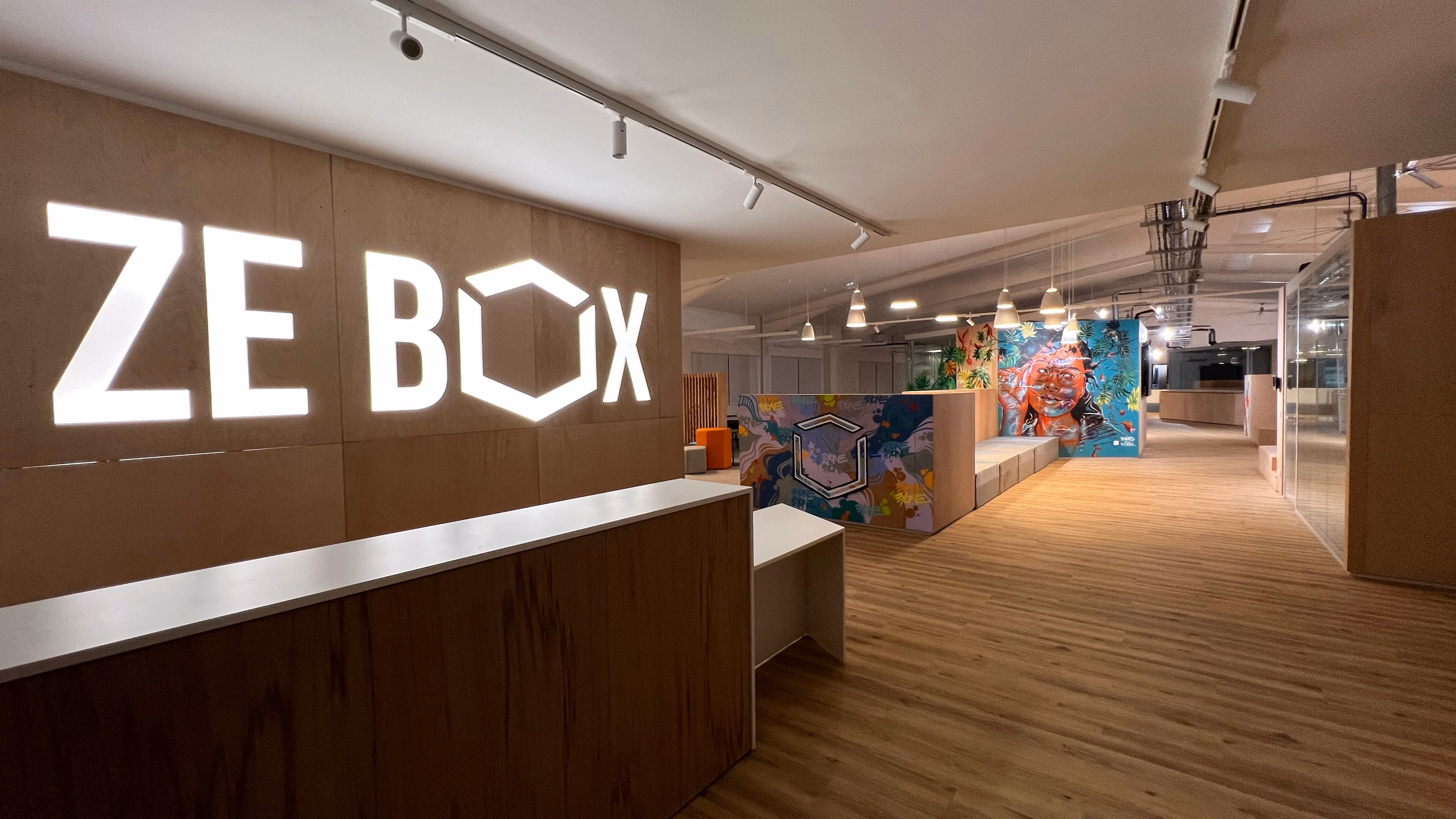 Reception area of Zebox Guadeloupe, an illuminated ZE BOX logo on a timber wall above a wooden desk, with colourful murals lining the corridor beyond