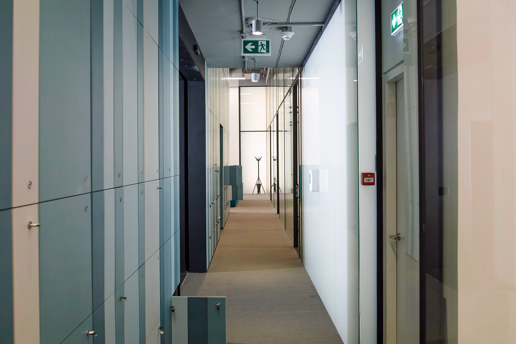 Corridor at Lot 114 in Beirut, lined with locker units on one side and frosted glass partitions on the other, leading to a bright open area