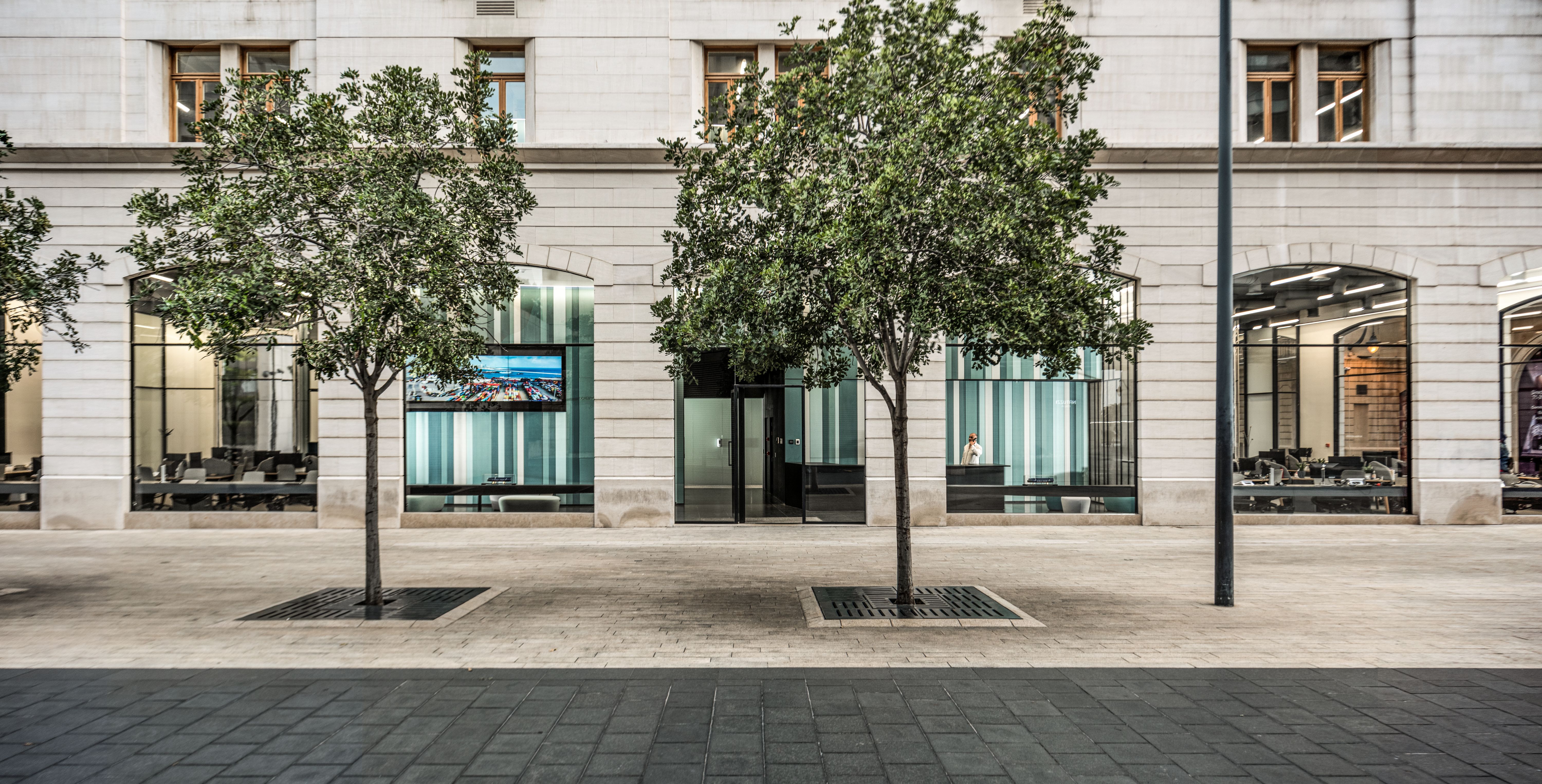 Ground floor storefront of Lot 114 in Beirut, arched stone facade with glazed offices behind two street trees on a paved plaza
