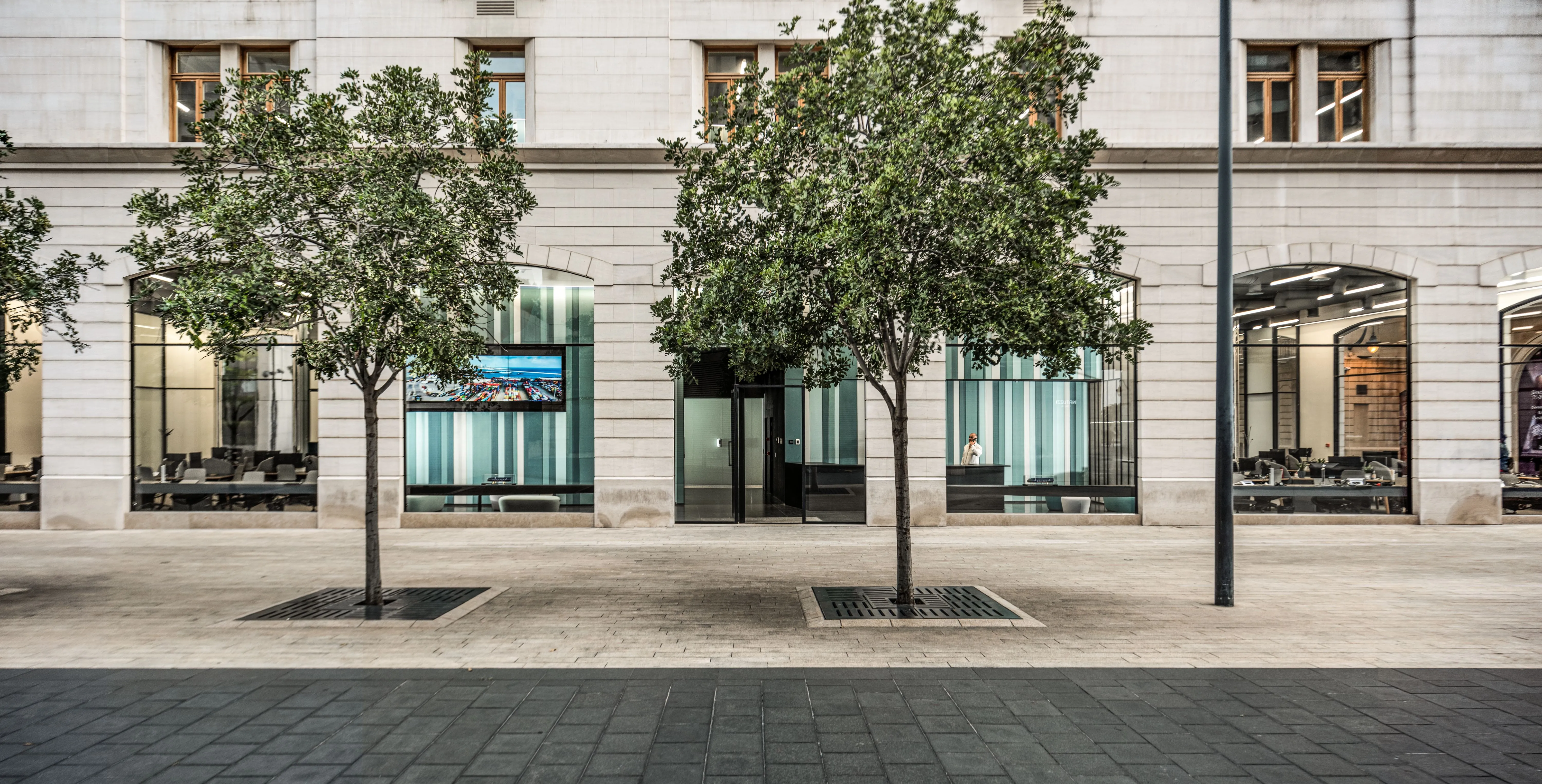 Ground floor storefront of Lot 114 in Beirut, arched stone facade with glazed offices behind two street trees on a paved plaza