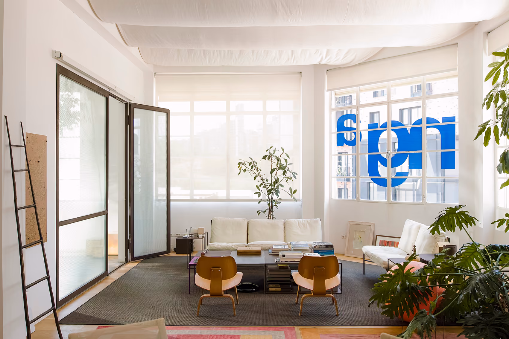 Lounge area at ngª Office in Beirut with white sofas, wooden chairs, potted plants and large windows bearing the ngª logo in reverse