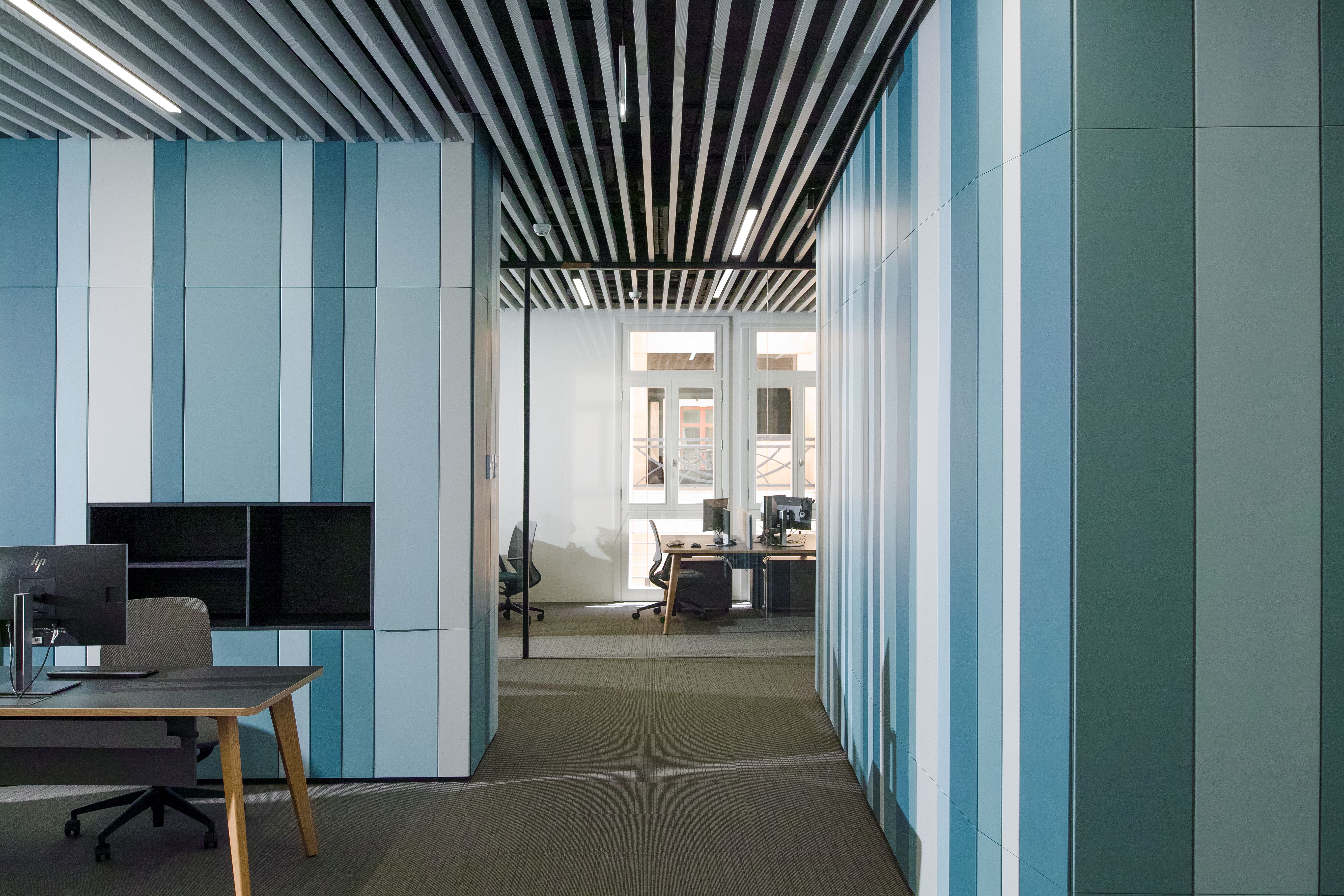 Open-plan office interior at Lot 114 in Beirut, curved striped blue wall panels, slatted ceiling, and workstations leading to a glazed meeting room