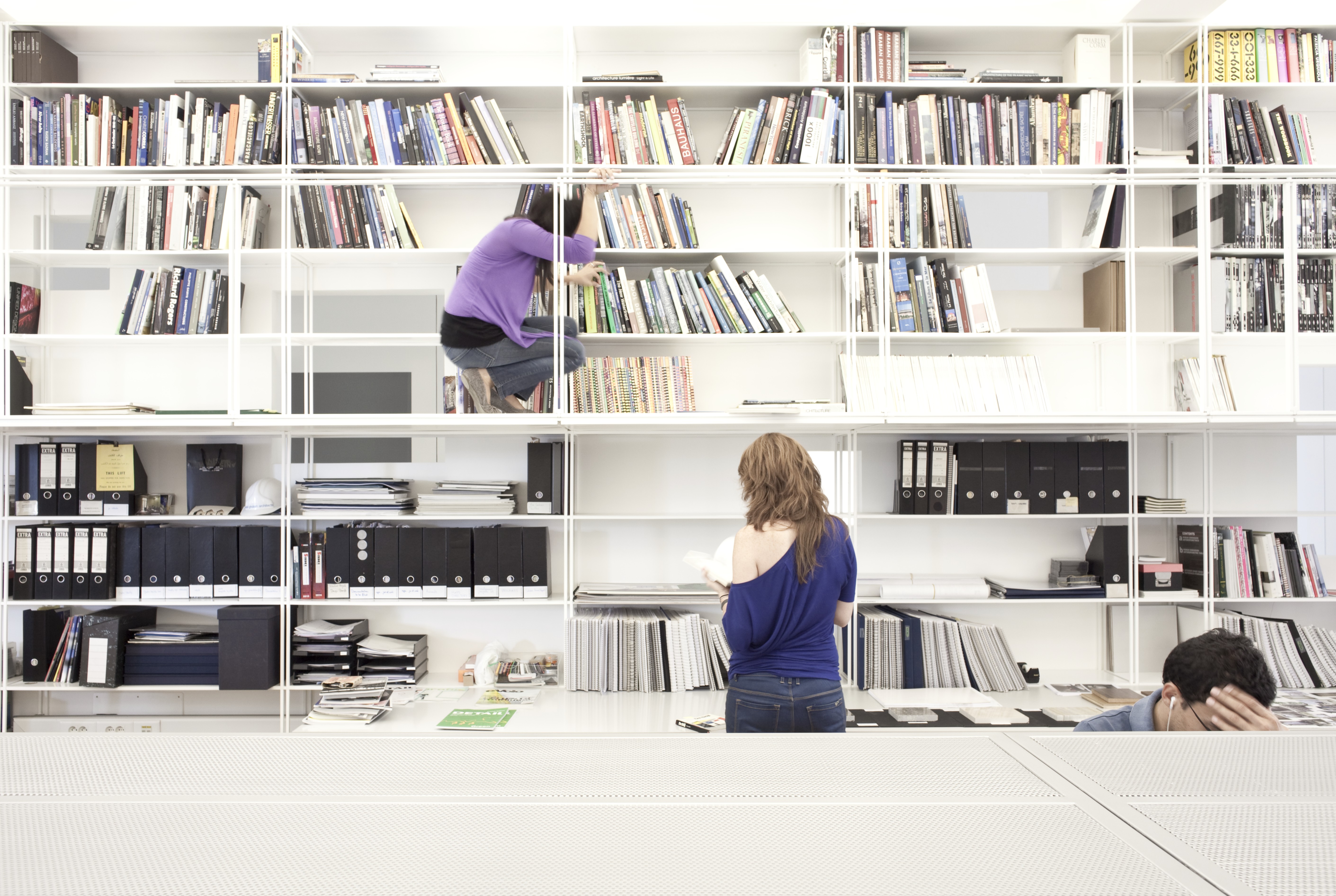 Staff browsing a white modular shelving wall of books and archive files at the ngª Office in Beirut, one perched on a ladder rung retrieving a title