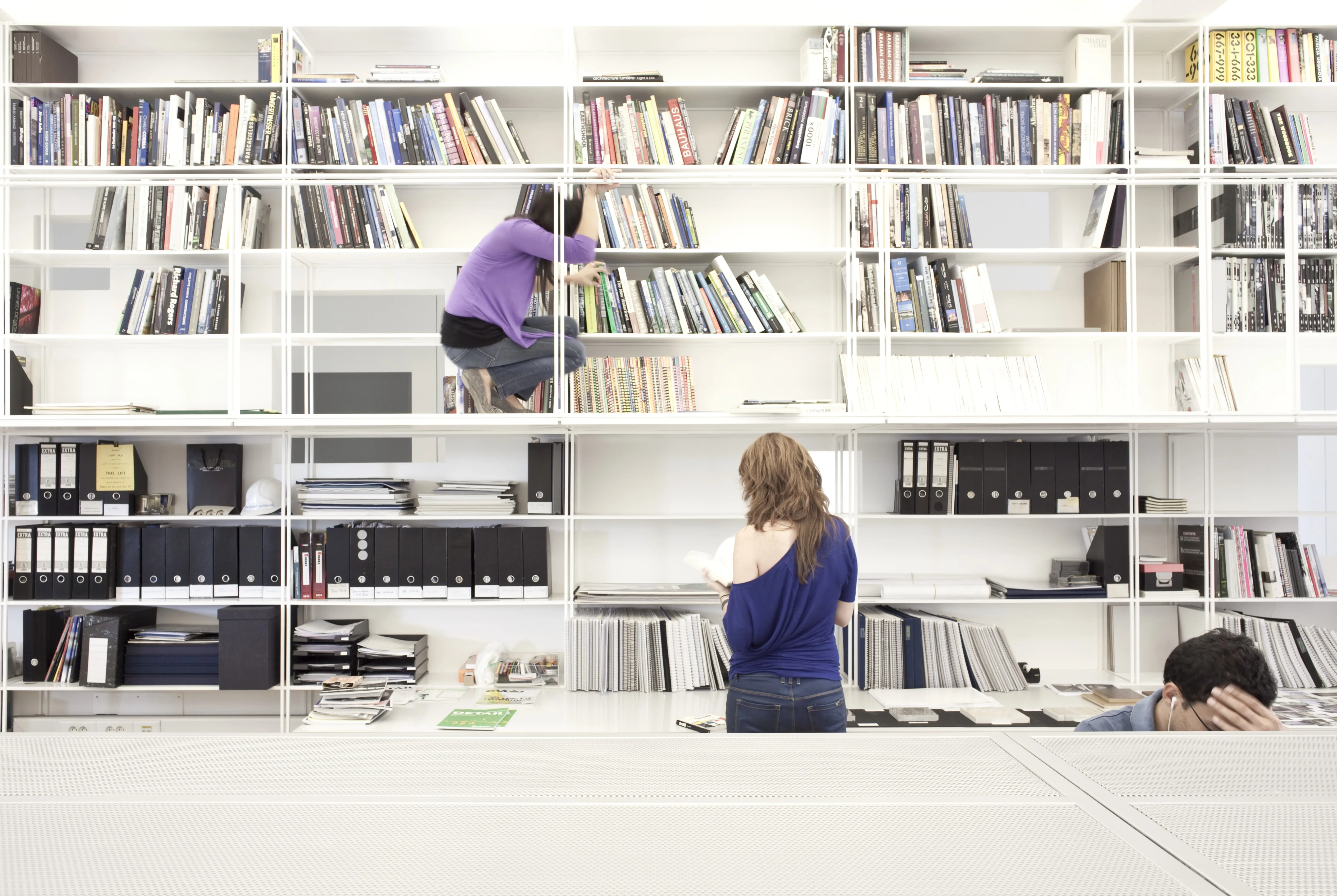 Staff browsing a white modular shelving wall of books and archive files at the ngª Office in Beirut, one perched on a ladder rung retrieving a title