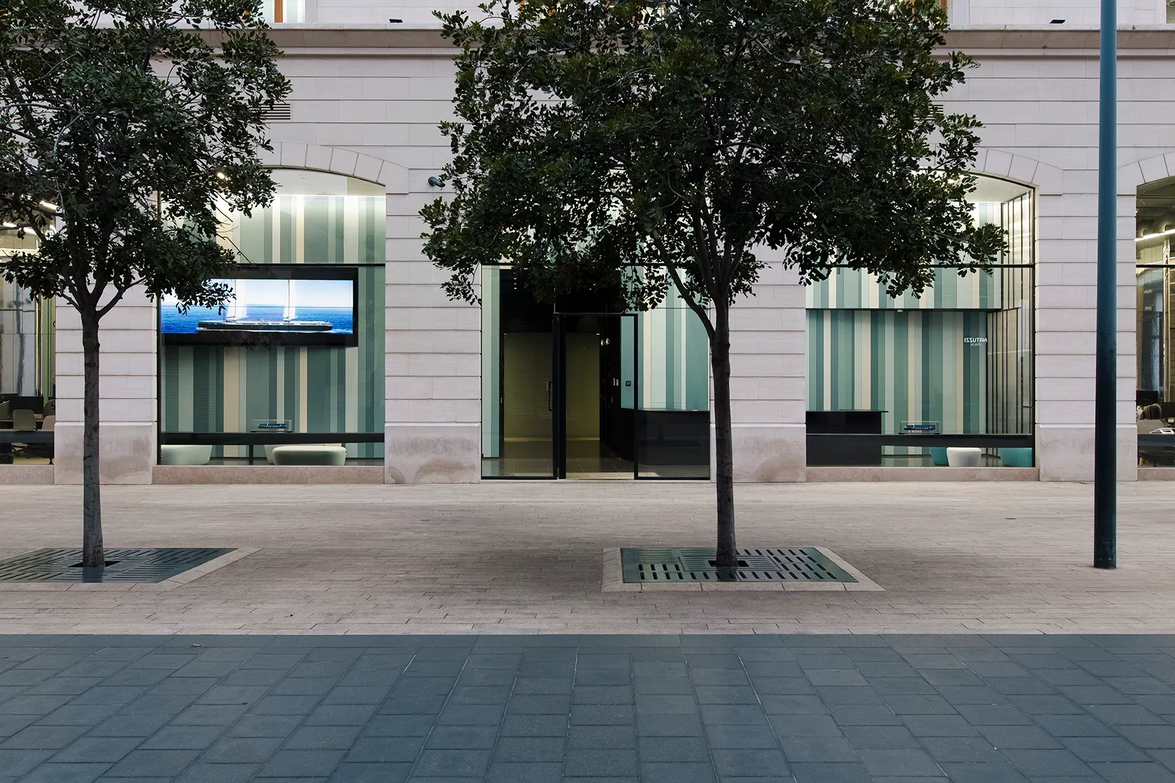 Ground floor storefronts of Lot 114 in Beirut, striped glass facades framed by trees along a paved pedestrian plaza