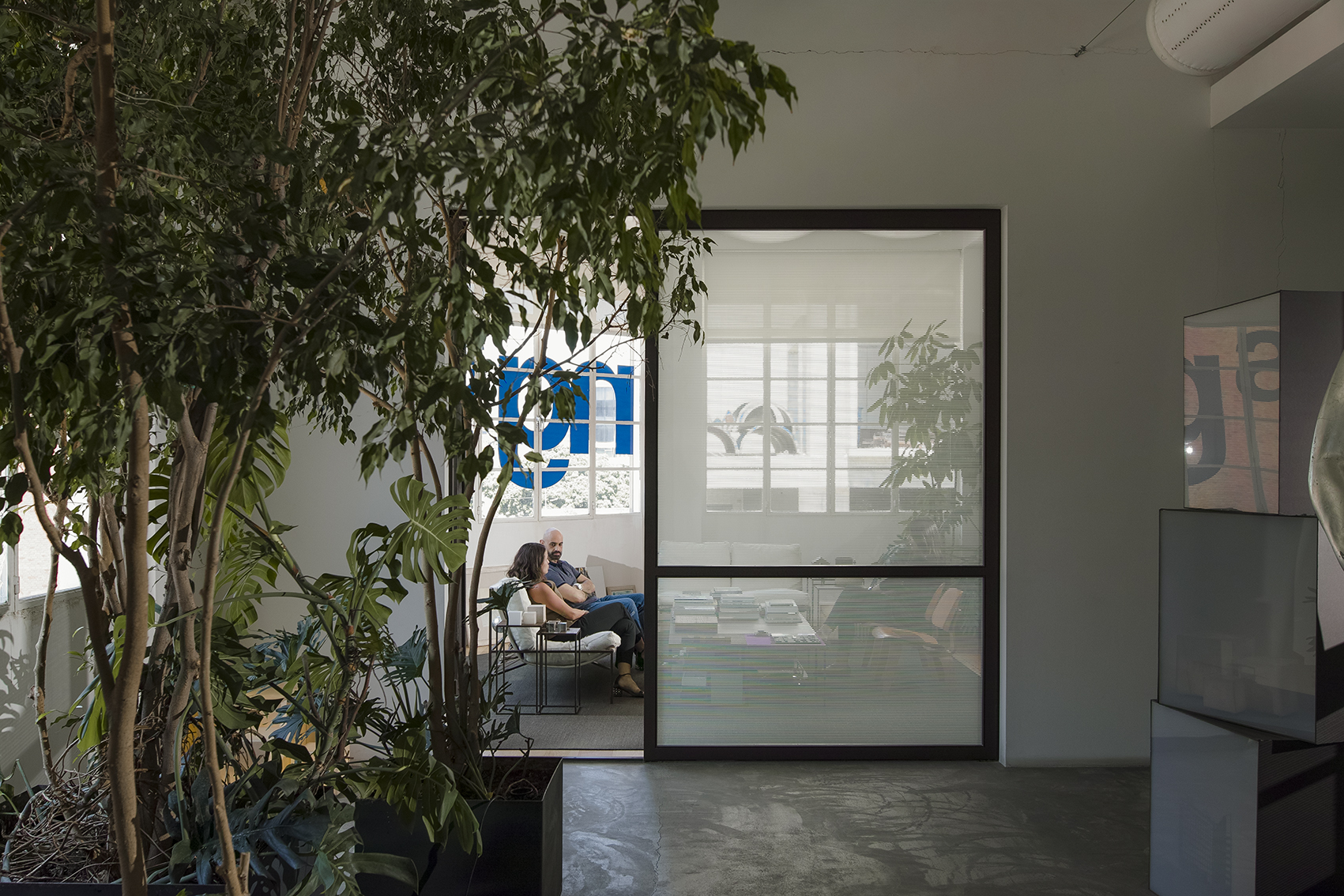 Lush indoor planting frames a glass-walled lounge at the ngª Office in Beirut, where two people sit talking beside a window with the firm's logo visible