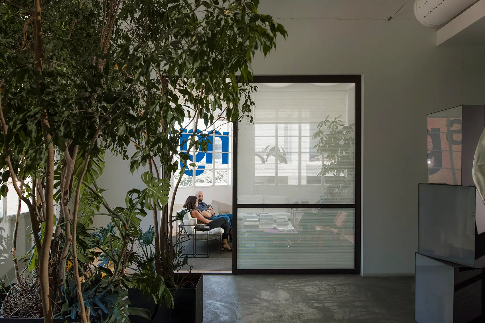 Lush indoor planting frames a glass-walled lounge at the ngª Office in Beirut, where two people sit talking beside a window with the firm's logo visible