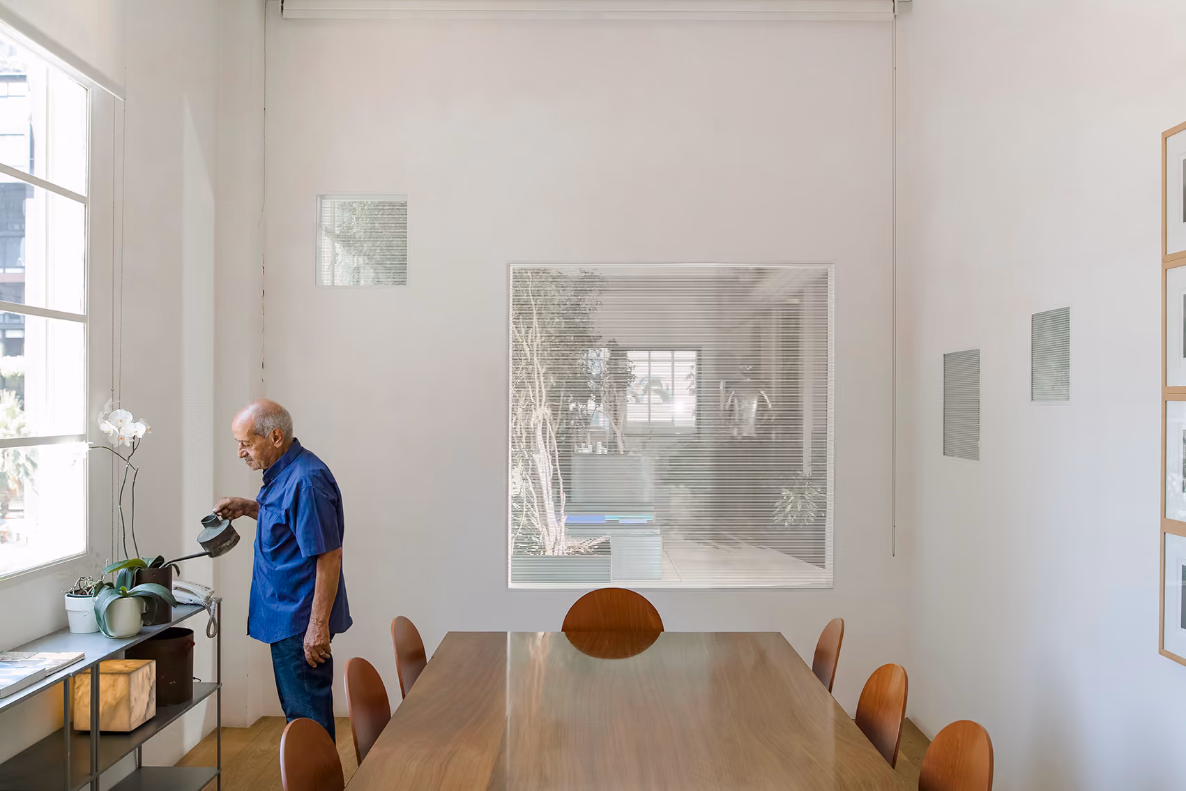 Man watering plants beside a long wooden meeting table in the ngª Office in Beirut, with an internal planted patio visible through an interior window