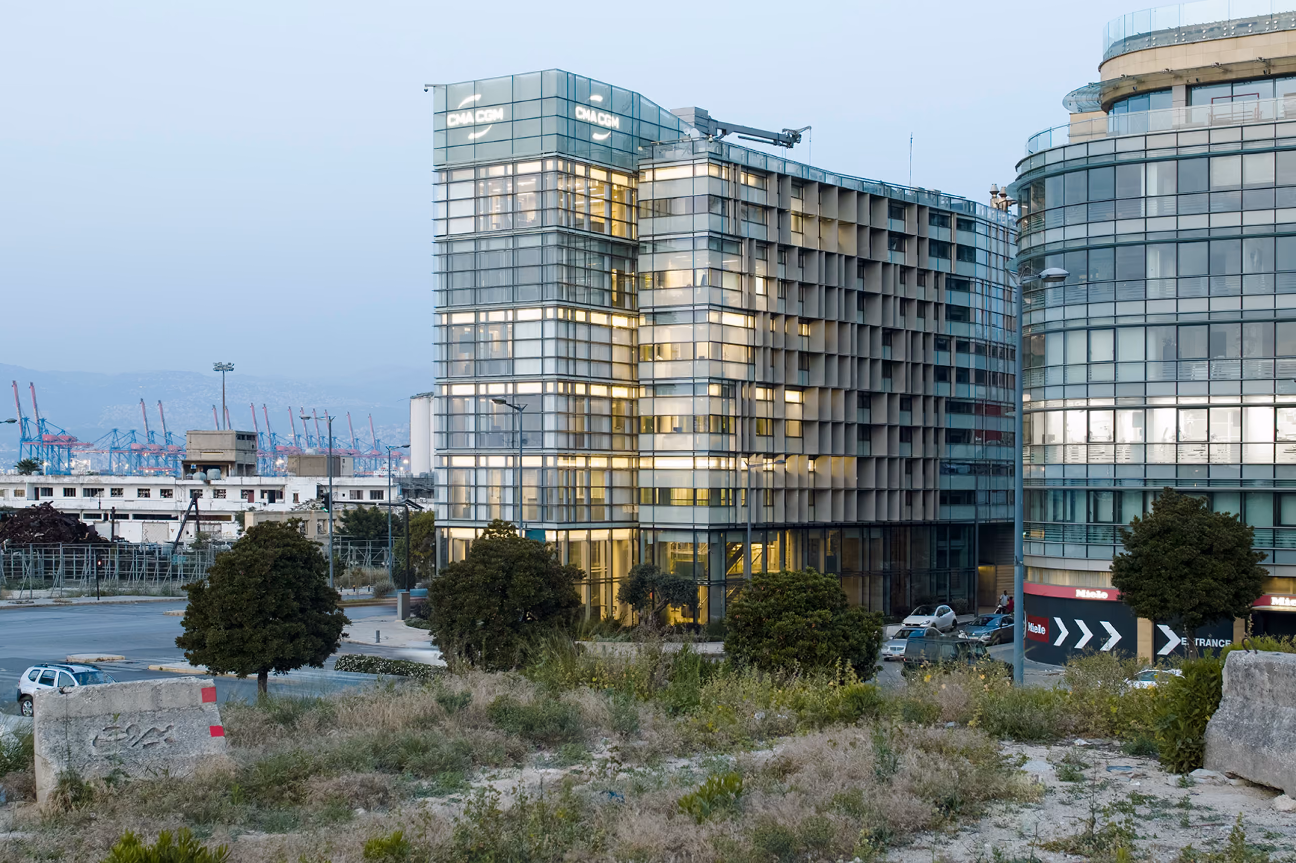 Twilight view of the refurbished CMA-CGM Headquarters in Beirut, its illuminated glass facade and timber brise-soleil rising near port cranes and shrubby foreground land